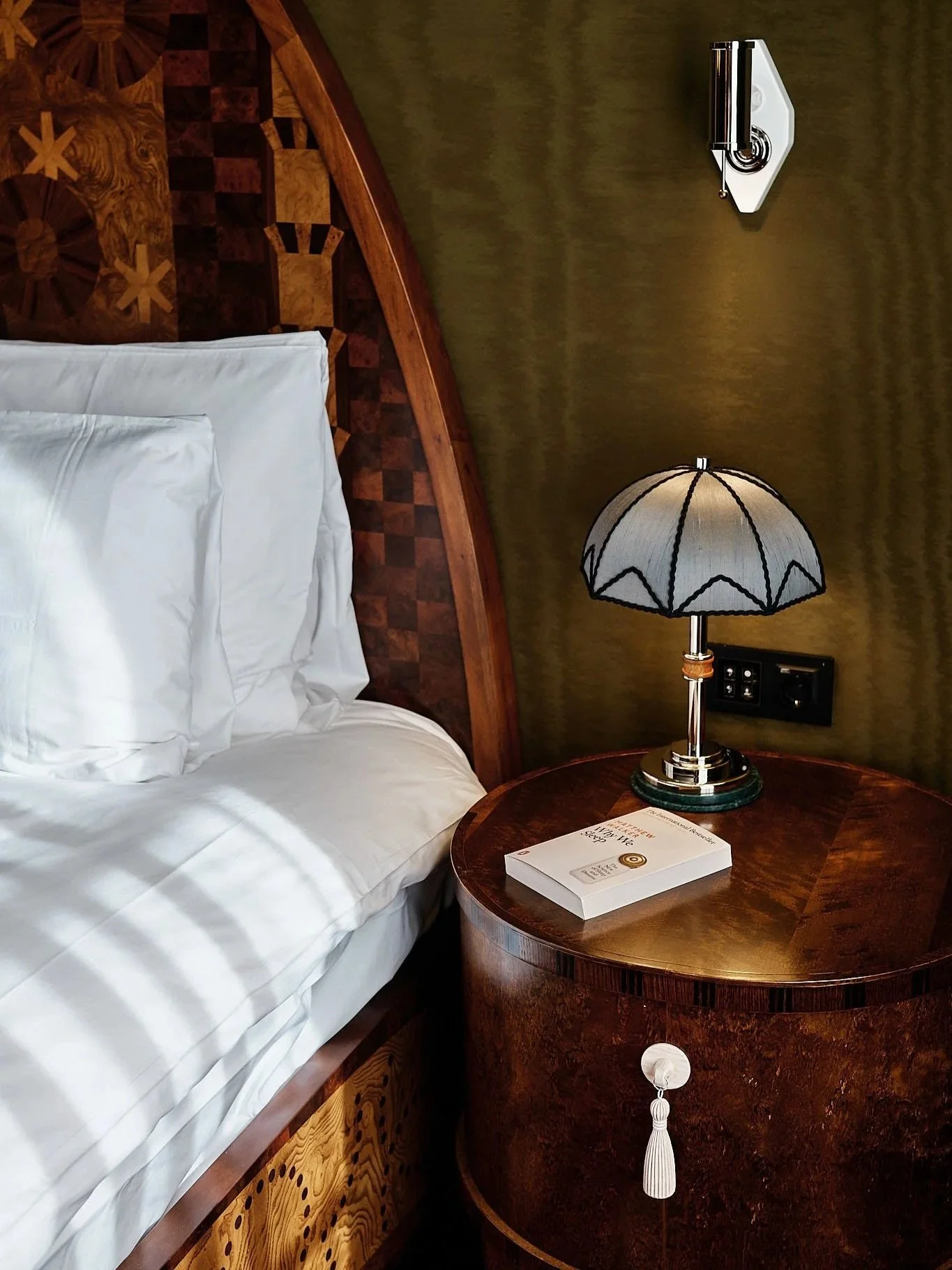 A hotel room corner showing part of a bed with white linens, a wooden headboard, a round wooden bedside table with a book and a vintage-style table lamp with a fabric shade, a green wall with a wall-mounted reading light, and electrical outlets.