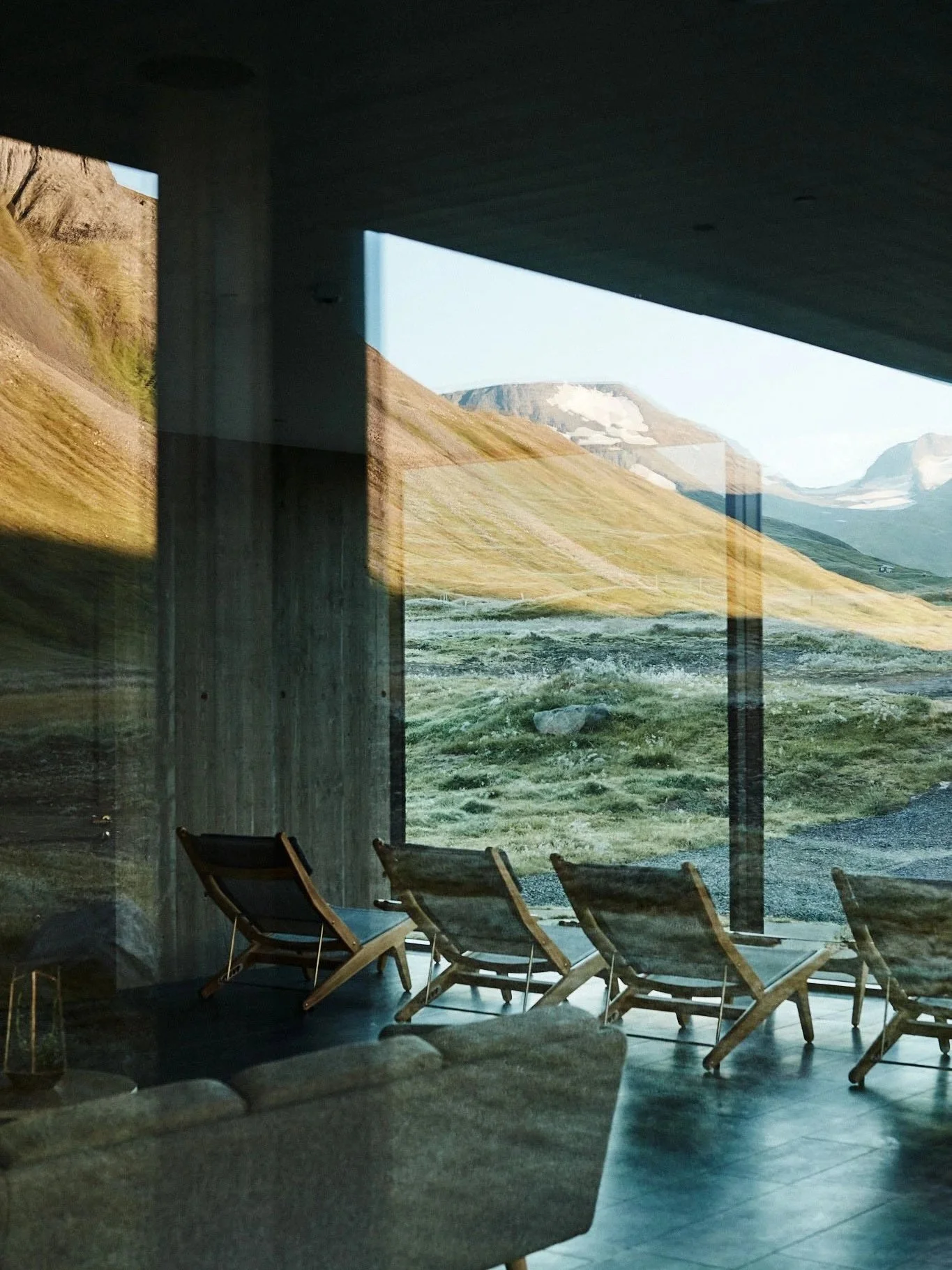View through large glass windows showing mountains, grassy terrain, and snow patches. Interior has lounge chairs and a sofa, with reflections on the glass.