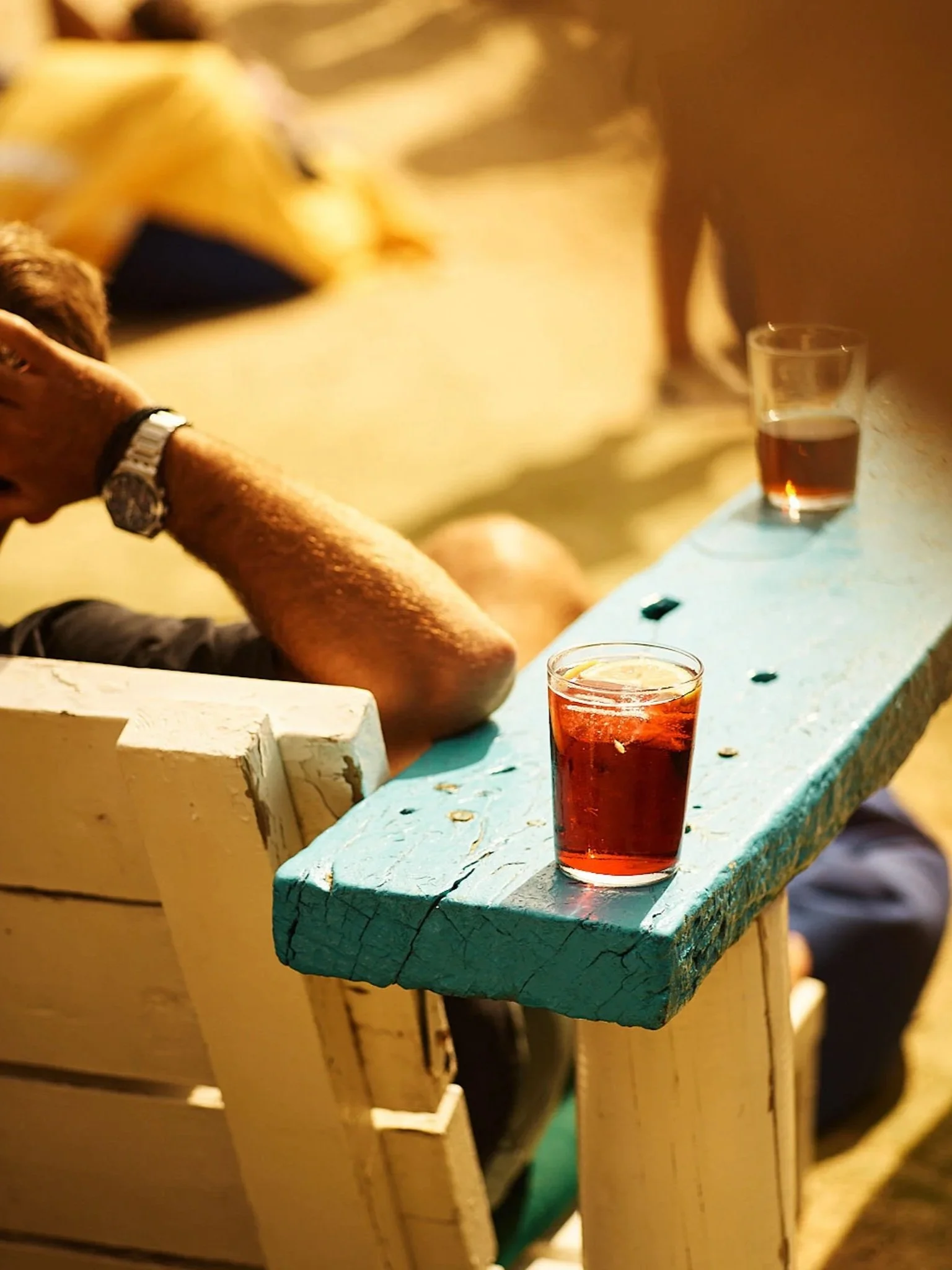 Two glasses of dark-colored beverages on a blue wooden bench, with a person sitting nearby resting their head on their hand, wearing a watch, in an outdoor setting.