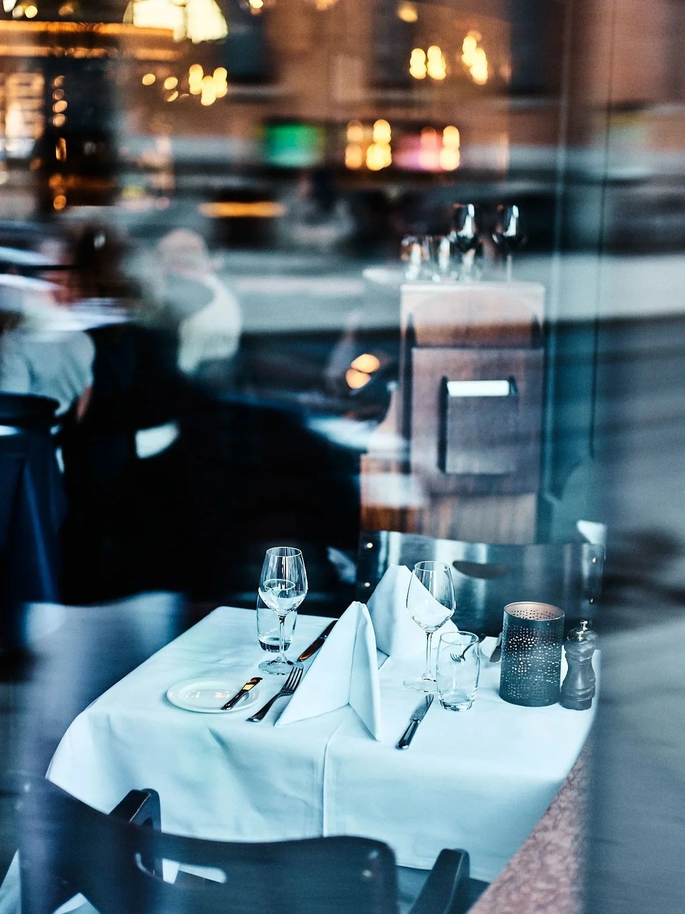 Empty restaurant table with white tablecloth, wine glasses, and folded napkins, viewed through a glass window.