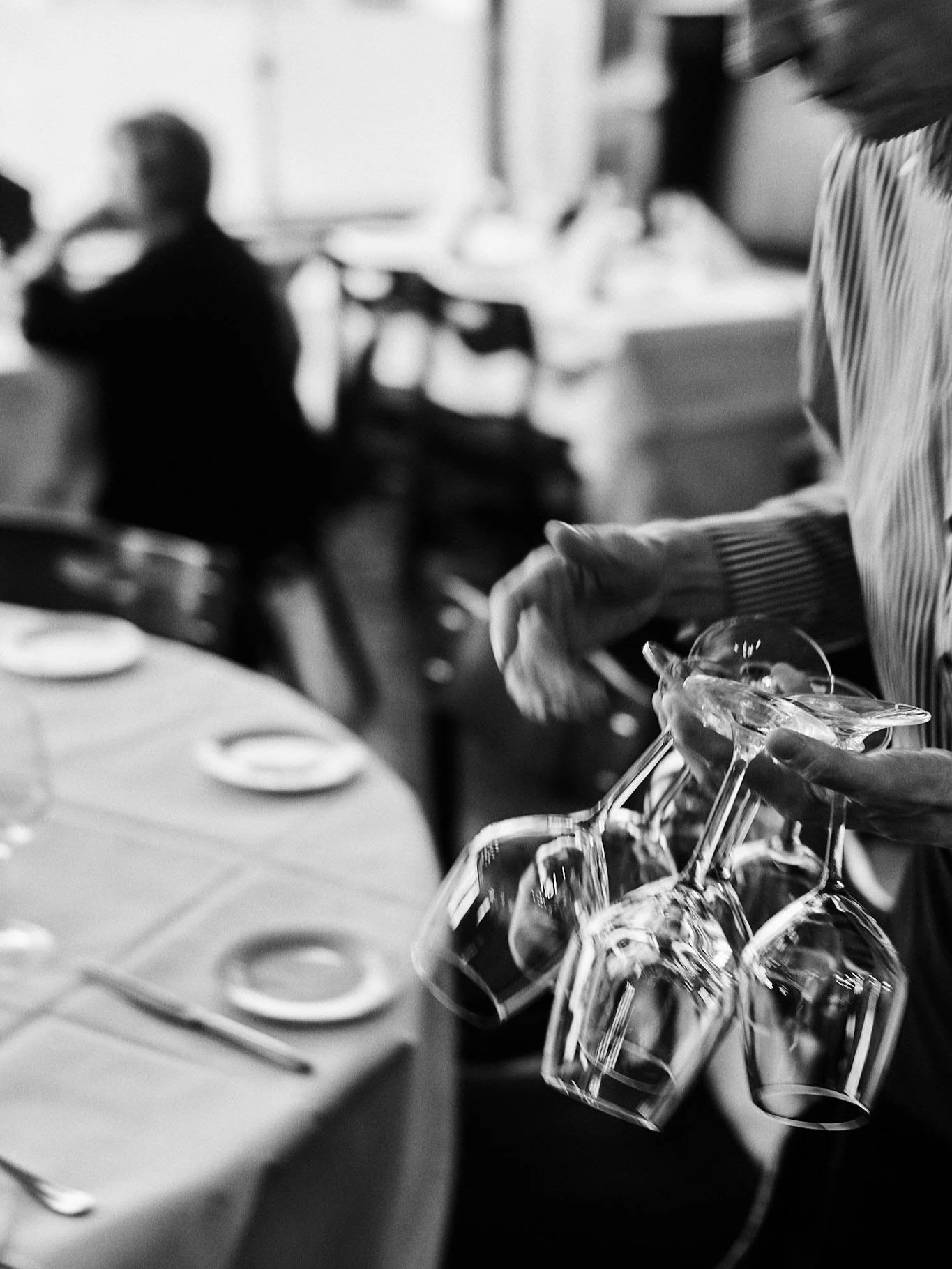 Person in a striped shirt holding several upside-down wine glasses in a restaurant setting.