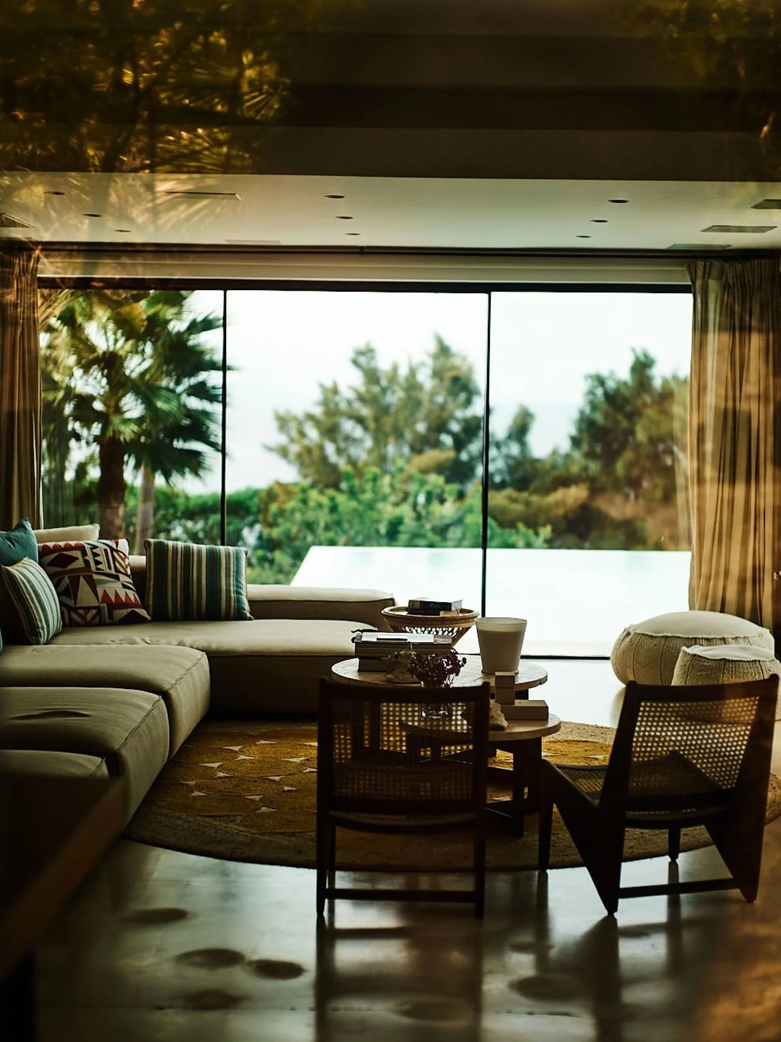 Living room with a large sectional sofa decorated with colorful pillows, a round coffee table with books and a white vase, and a view of trees through large glass sliding doors.