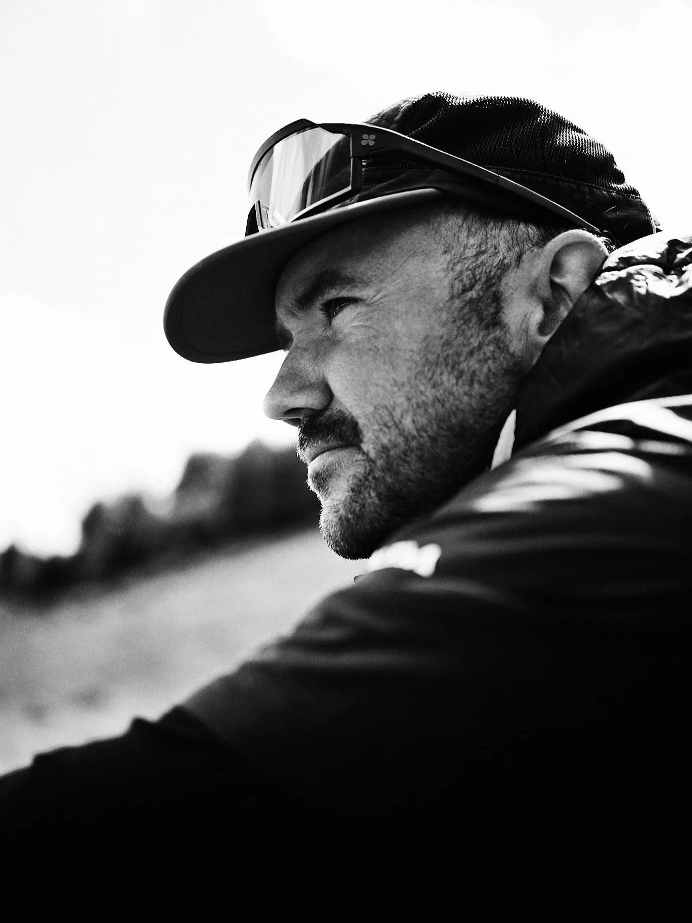 A black and white close-up photo of a man wearing a cap and sunglasses, looking to the left with a serious expression.