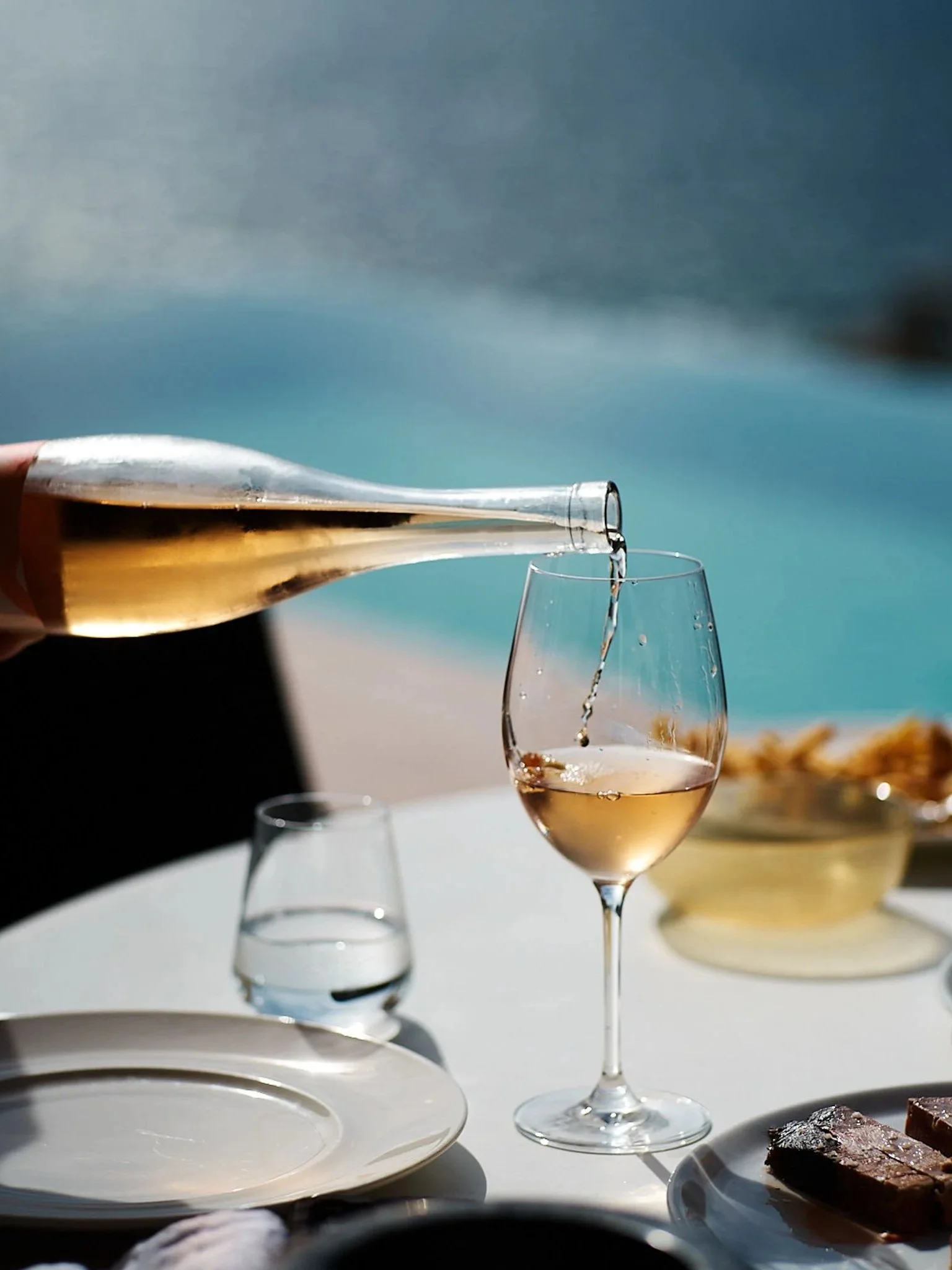 A hand pouring rosé wine into a glass on a white table near a body of water, with plates of food and a bowl in the background.