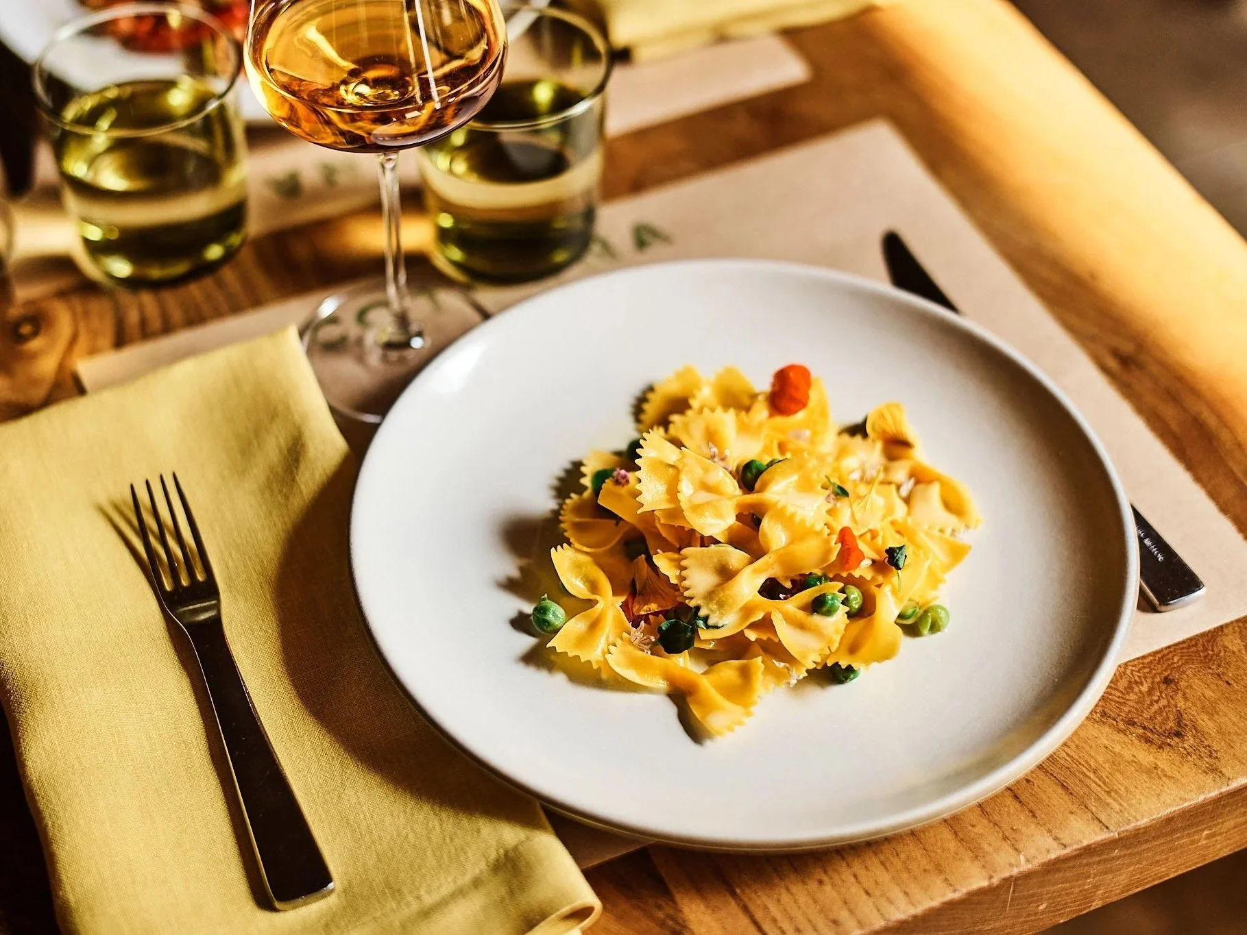 A white plate of farfalle pasta with green peas and small orange garnishes on a wooden table. There is a fork with a black handle on a yellow cloth napkin to the left, and a knife on a beige placemat to the right. In the background, there are glasses