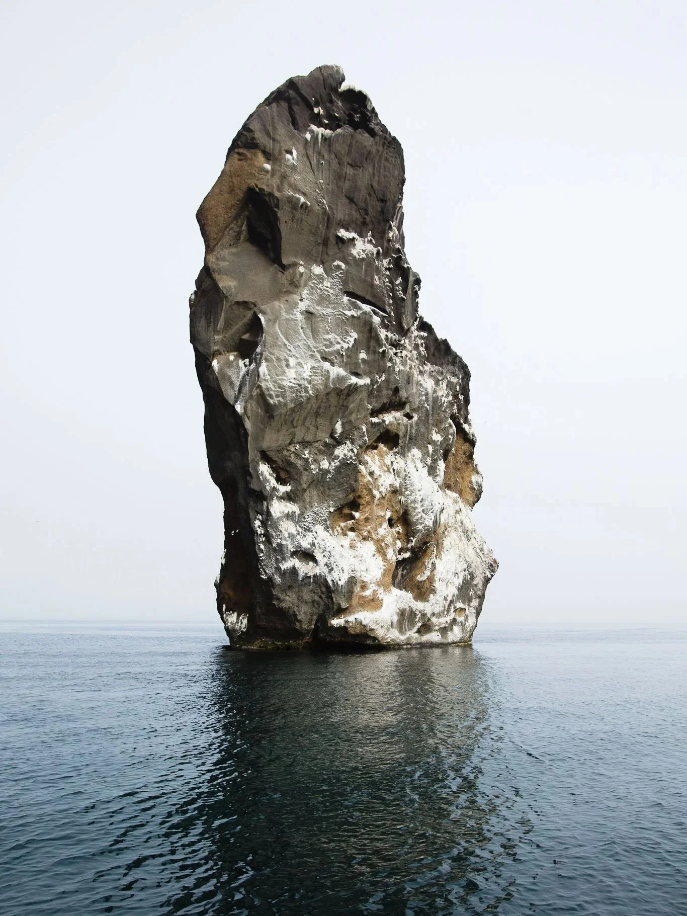 A large, tall rock formation rising out of the water, with a smooth, rounded base and a rugged, uneven top, under a pale sky.