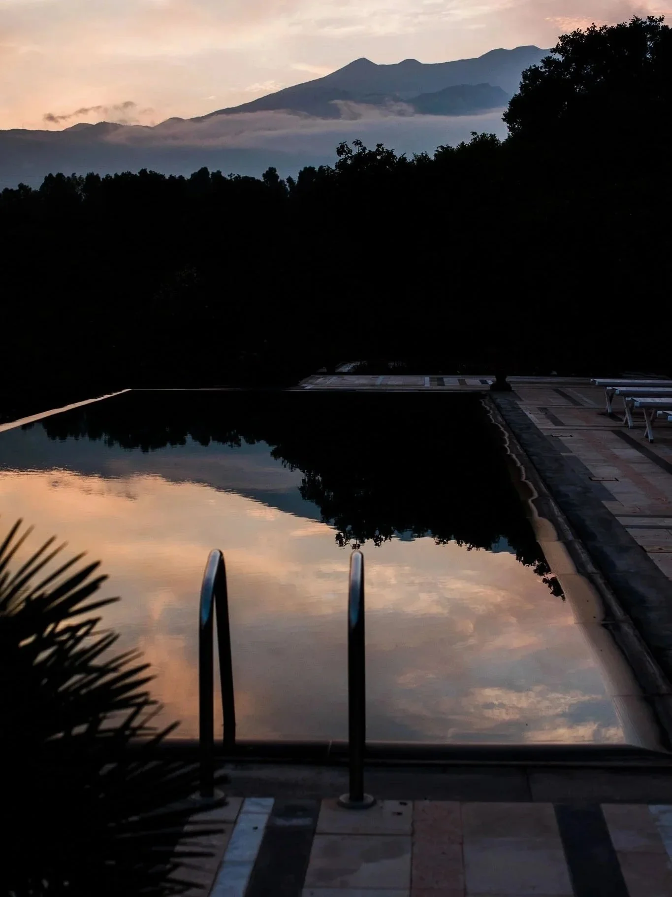 Sunset view of a mountain range with clouds, reflected in the still water of a swimming pool, surrounded by trees and a tiled poolside area.
