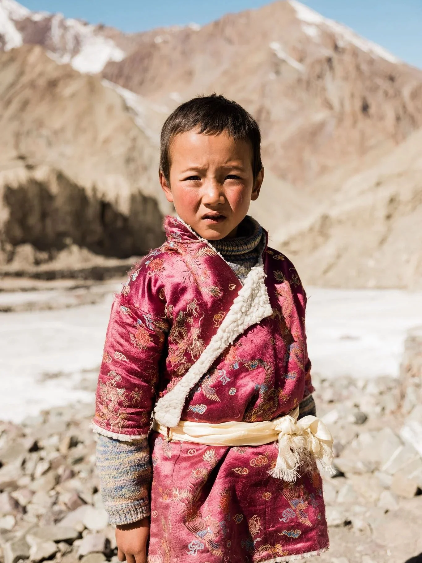 Young boy standing outdoors in a mountainous, rocky landscape with snow patches, wearing traditional clothing, looking at the camera with a serious expression.