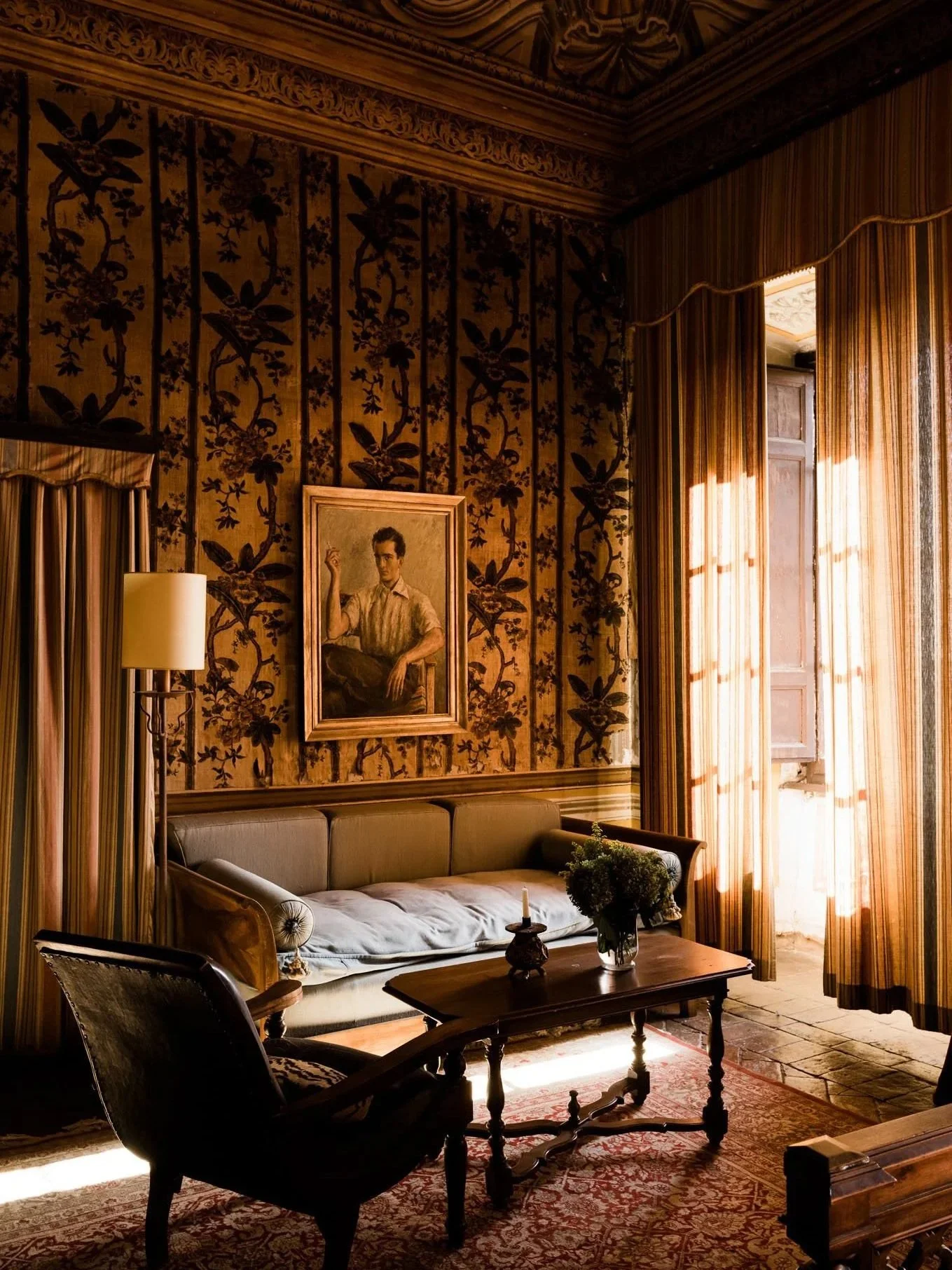 A vintage living room with a beige sofa, an armchair, a wooden coffee table with a vase of flowers, patterned curtains, an ornate carpet, a portrait on the wall, and sunlight filtering through large windows.