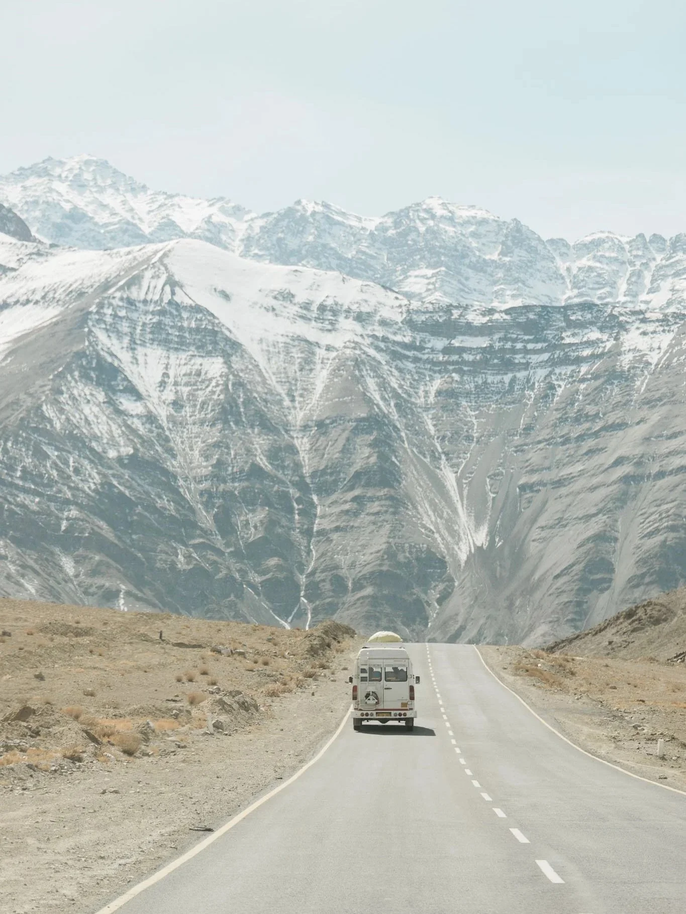 A white camper van driving on a winding mountain road with snow-capped peaks in the background.