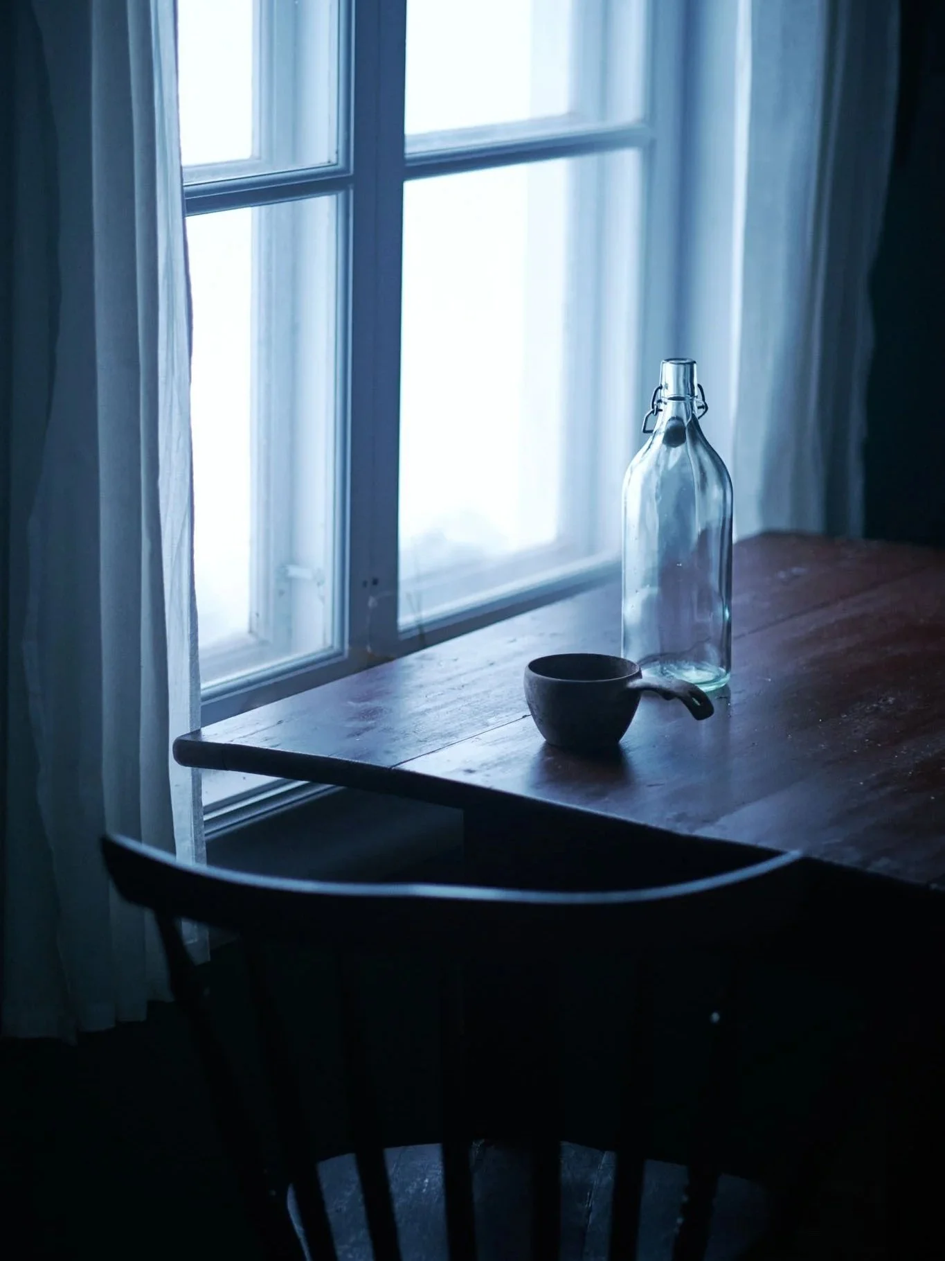 A wooden table with a glass bottle and a ceramic cup near a window with sheer curtains, in a softly lit room.