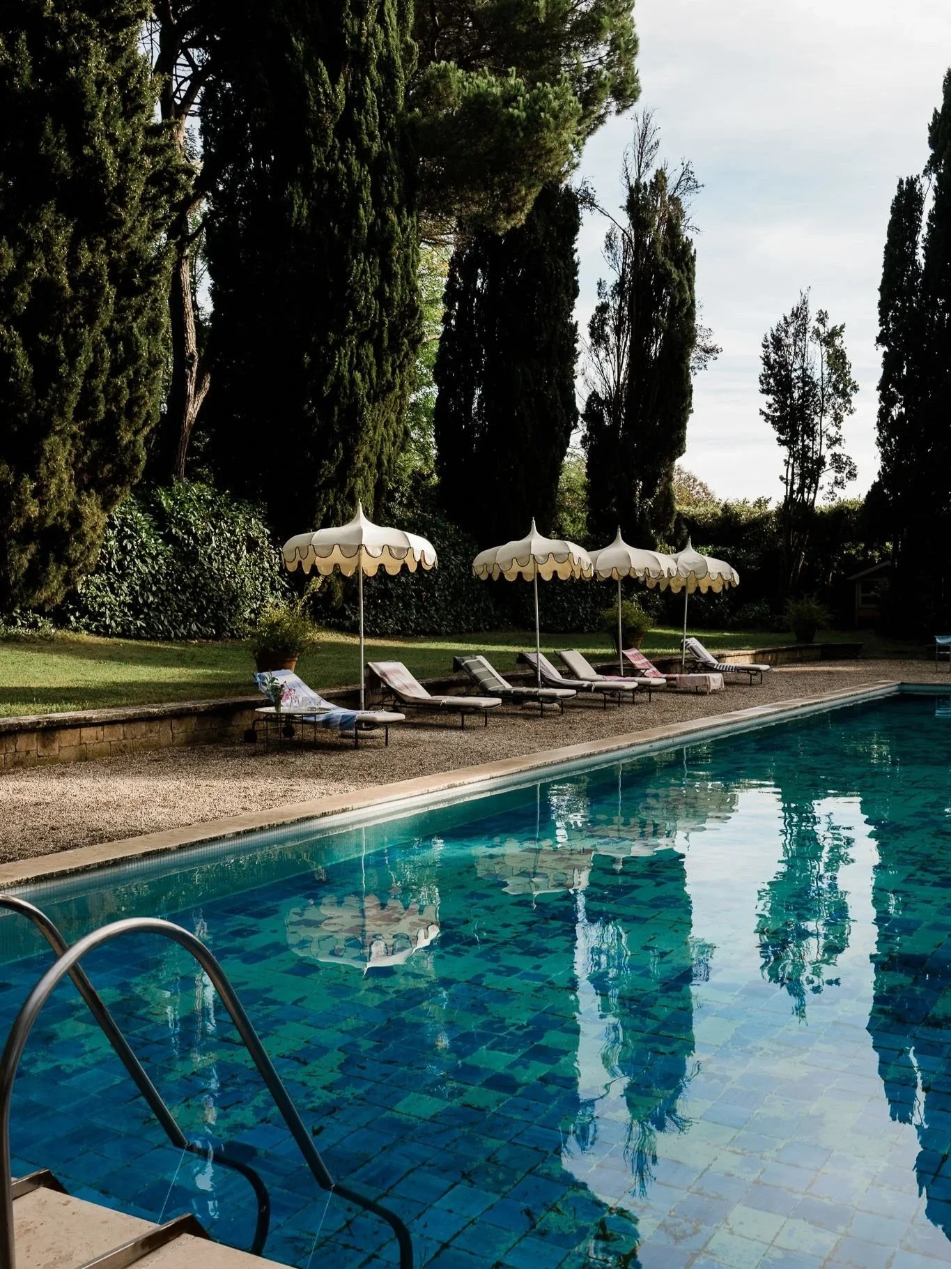 A swimming pool with blue tiles, surrounded by lounge chairs, umbrellas, and tall trees in the background.