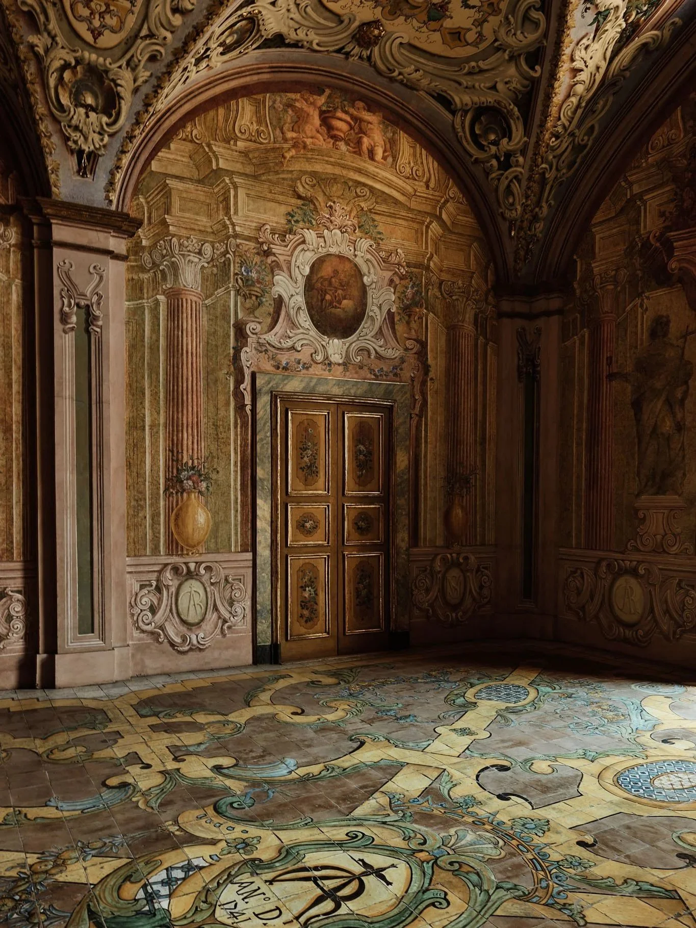 An ornate, baroque-style room with detailed carvings on the walls and ceiling, featuring frescoes, columns, and a wooden door with floral designs. The floor is decorated with intricate, colorful tile patterns.