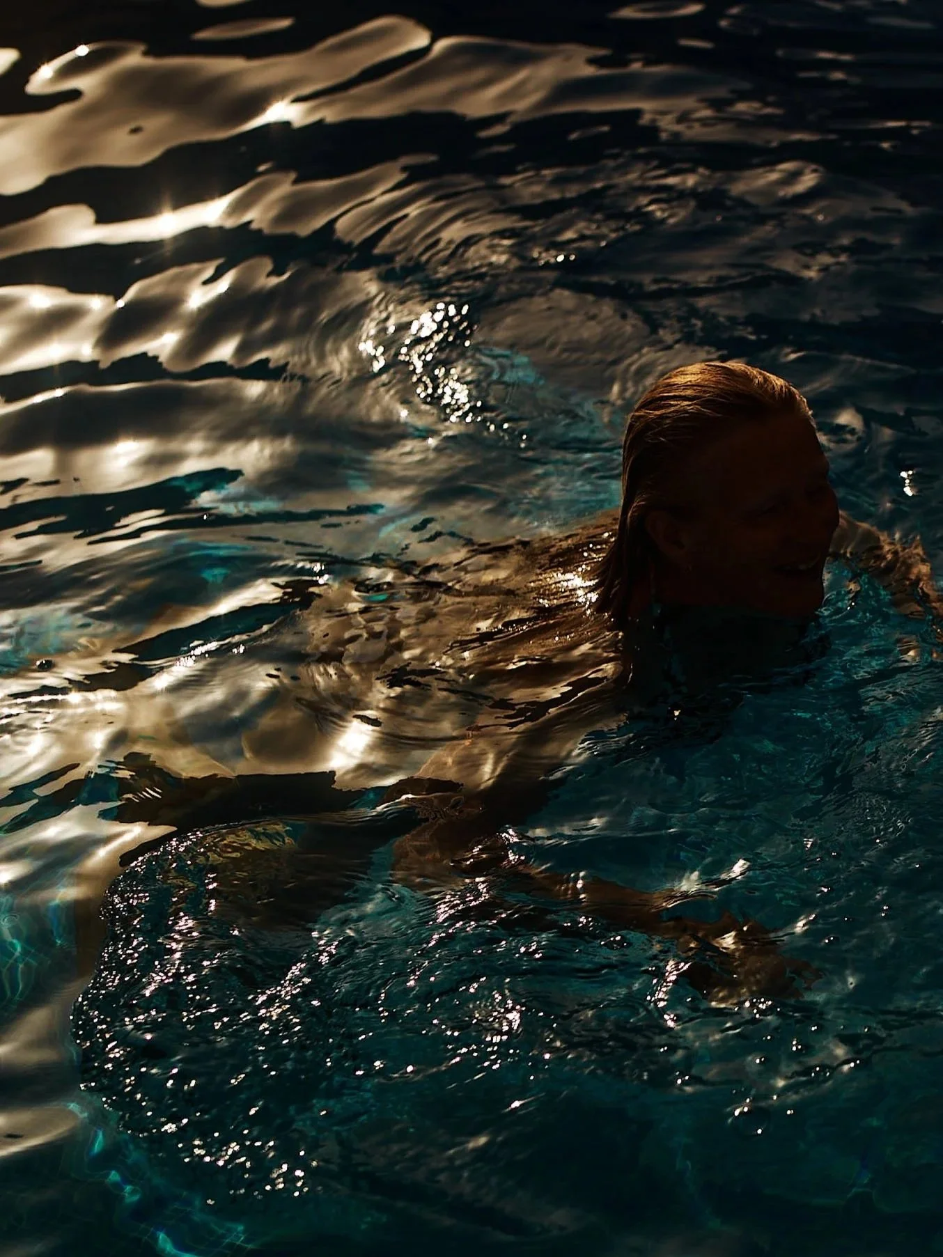 Person swimming in a pool at sunset or nighttime, with reflections on the water.