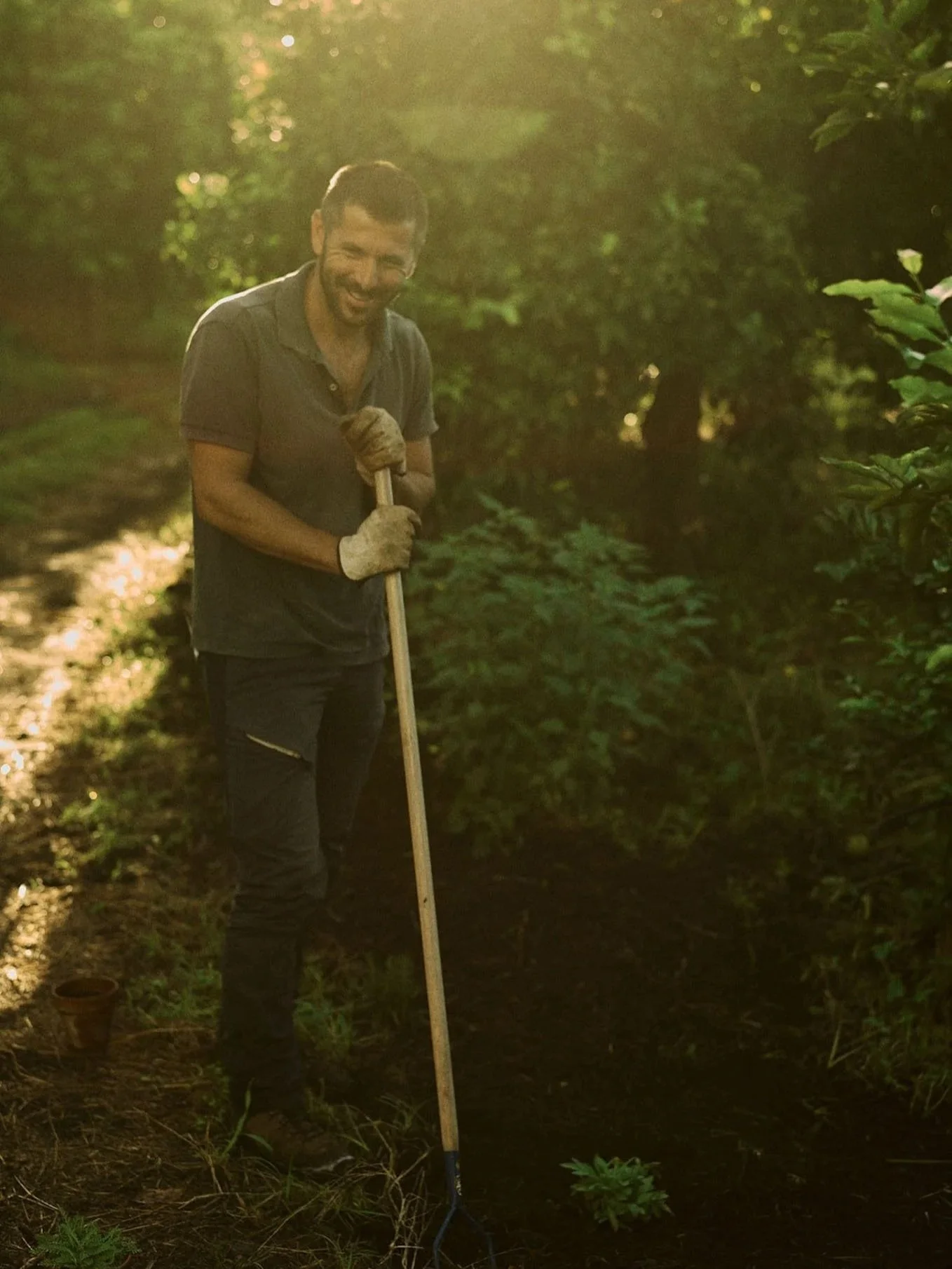 A man smiling while gardening outdoors in a wooded area, using a gardening fork. He is wearing gloves and dark clothing, with sunlight filtering through the trees.