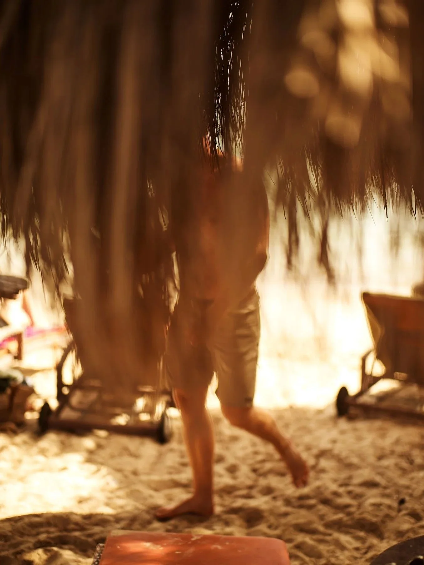 A person walking on a sandy beach, seen through a thatched roof in the foreground, with two chairs on the sand to the sides, during sunset or sunrise.