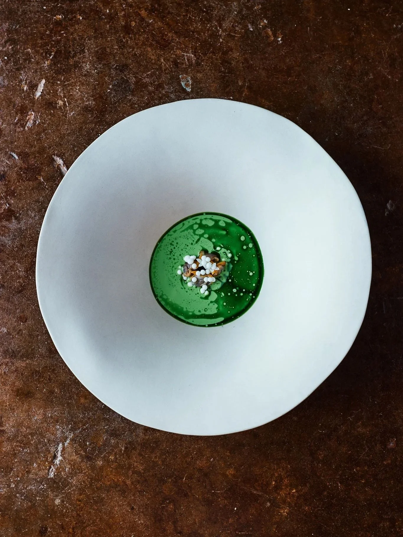 A small green dish with white and black toppings on a white plate, placed on a brown textured surface.