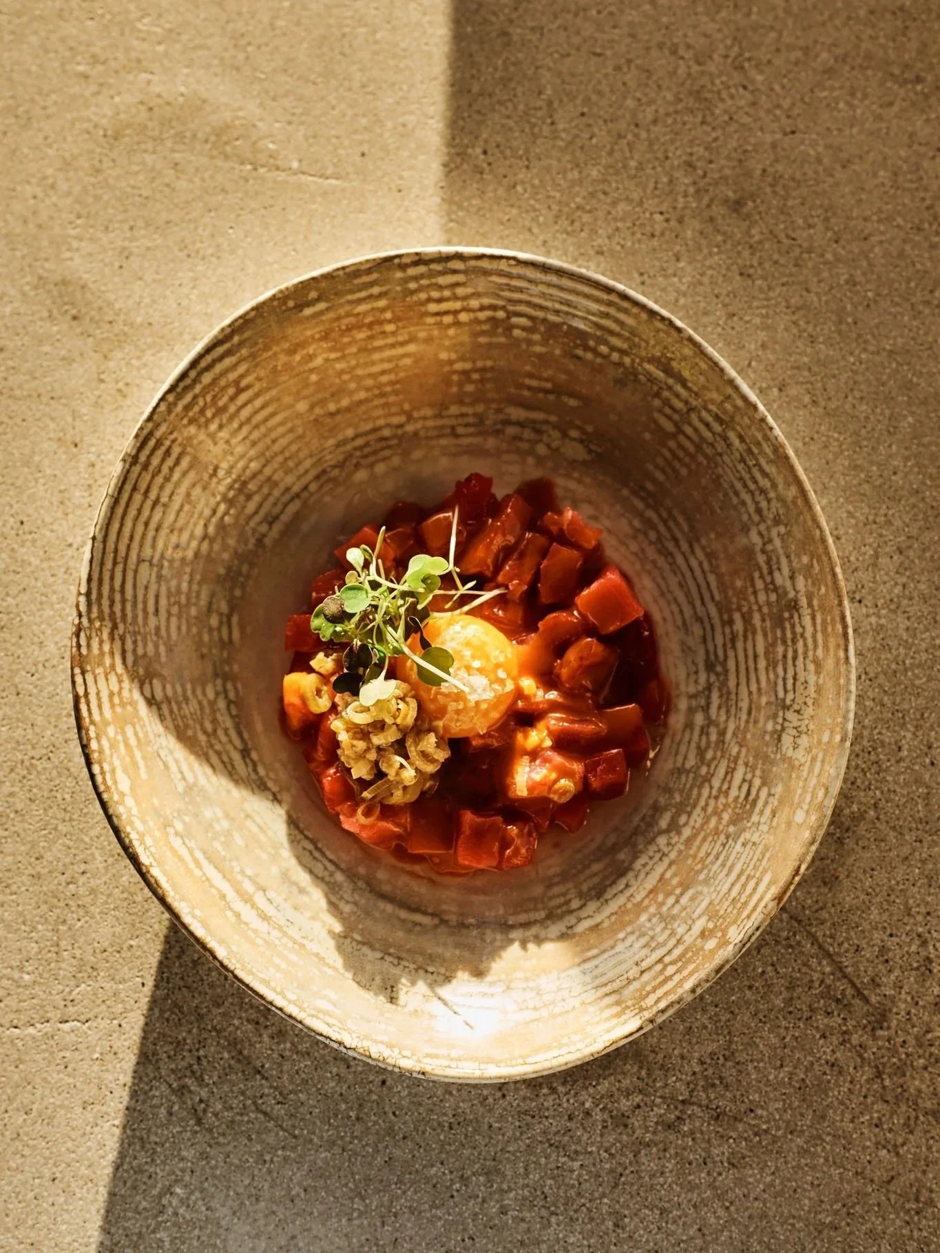 A bowl of diced raw meat, topped with microgreens, a poached egg, and a small cluster of grains, on a gray surface.