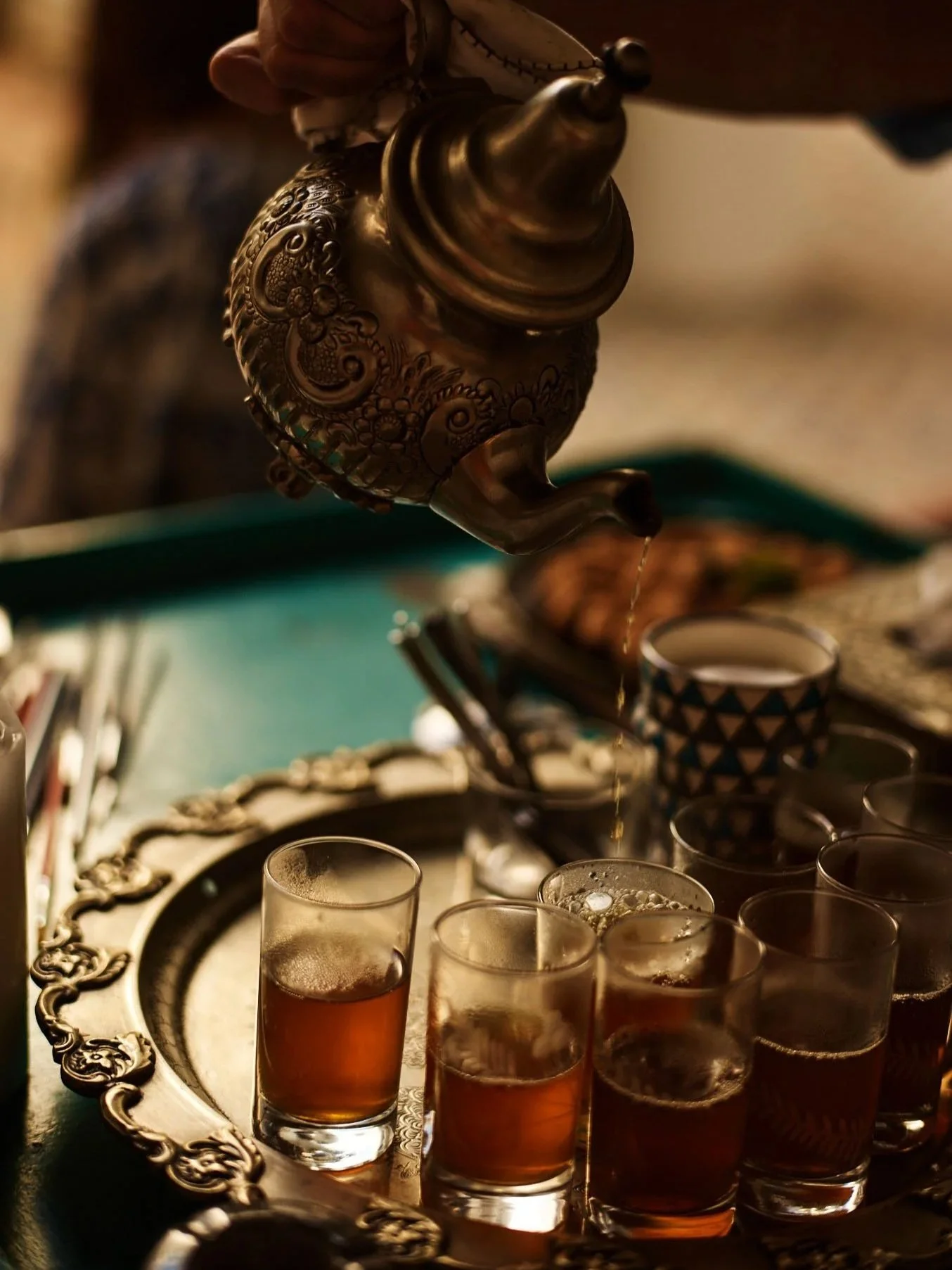 A decorative metal teapot pouring tea into small glasses on a silver tray.