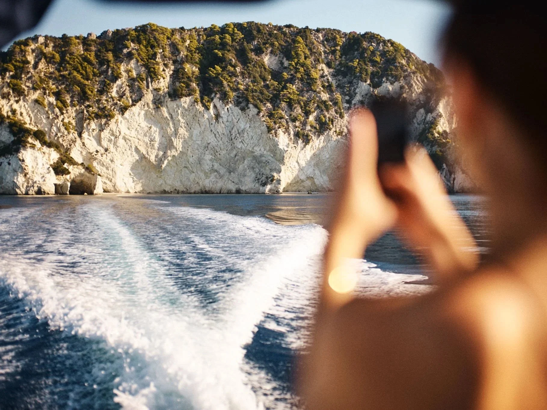 A person taking a photo with a camera on a boat, with a backdrop of rocky cliffs covered in greenery and the water leaving a wake behind.