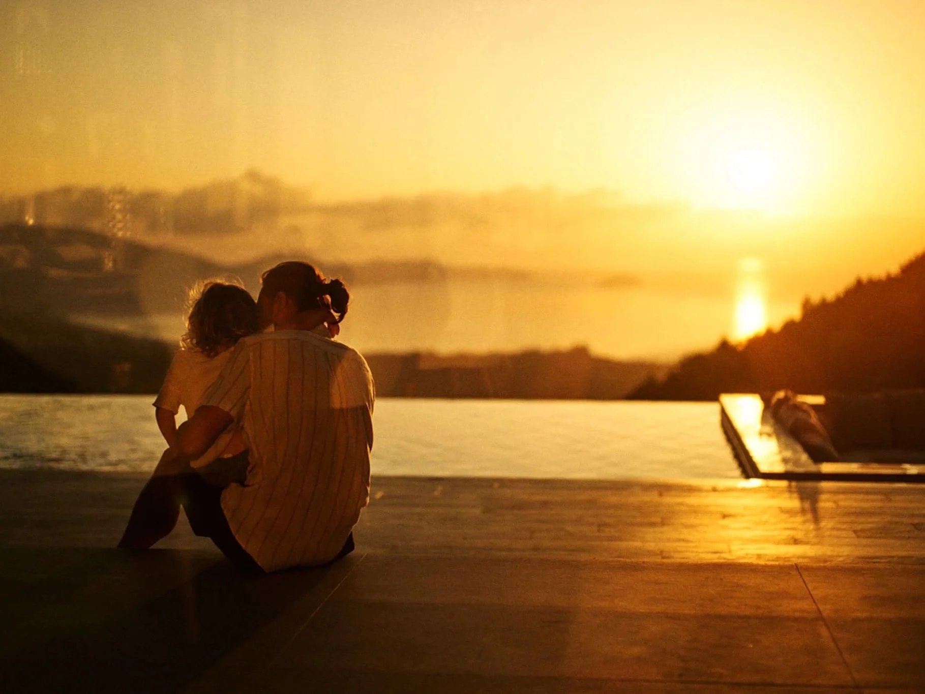 A woman and a young girl sit close together, sharing a kiss on a wooden deck at sunset with a scenic water and mountain view in the background.