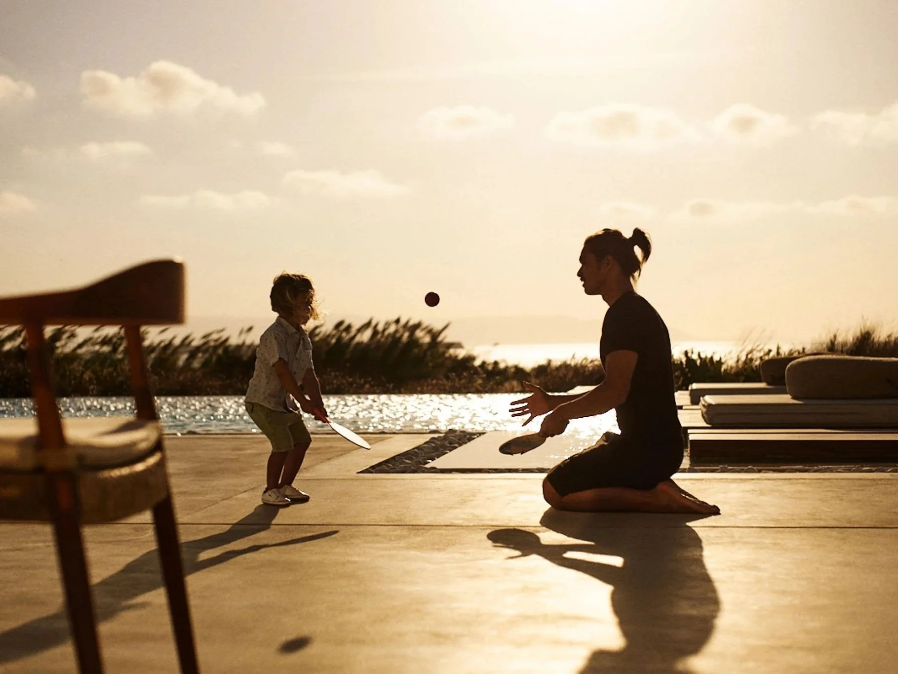 A woman kneeling on the ground playing with a young child at sunset near a pool, with the child holding a paddle and a ball in mid-air.