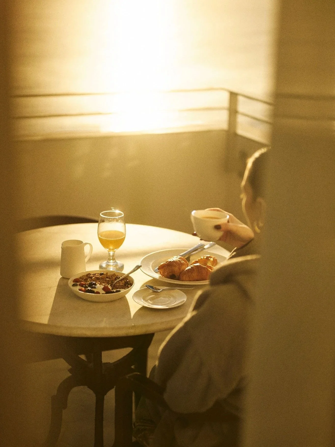 A woman sitting at a round table, seen from behind, enjoying breakfast with croissants, cereal, coffee, and a glass of juice, in a warmly lit room.