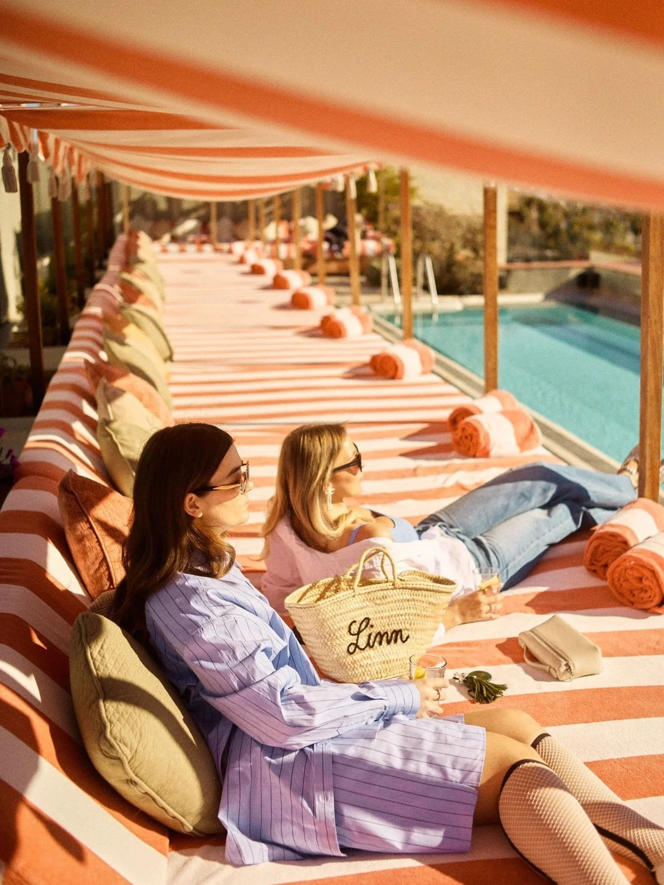 Two women relaxing on striped lounge chairs by a poolside, with rolled towels, pillows, and a straw bag, under a red and white striped canopy.
