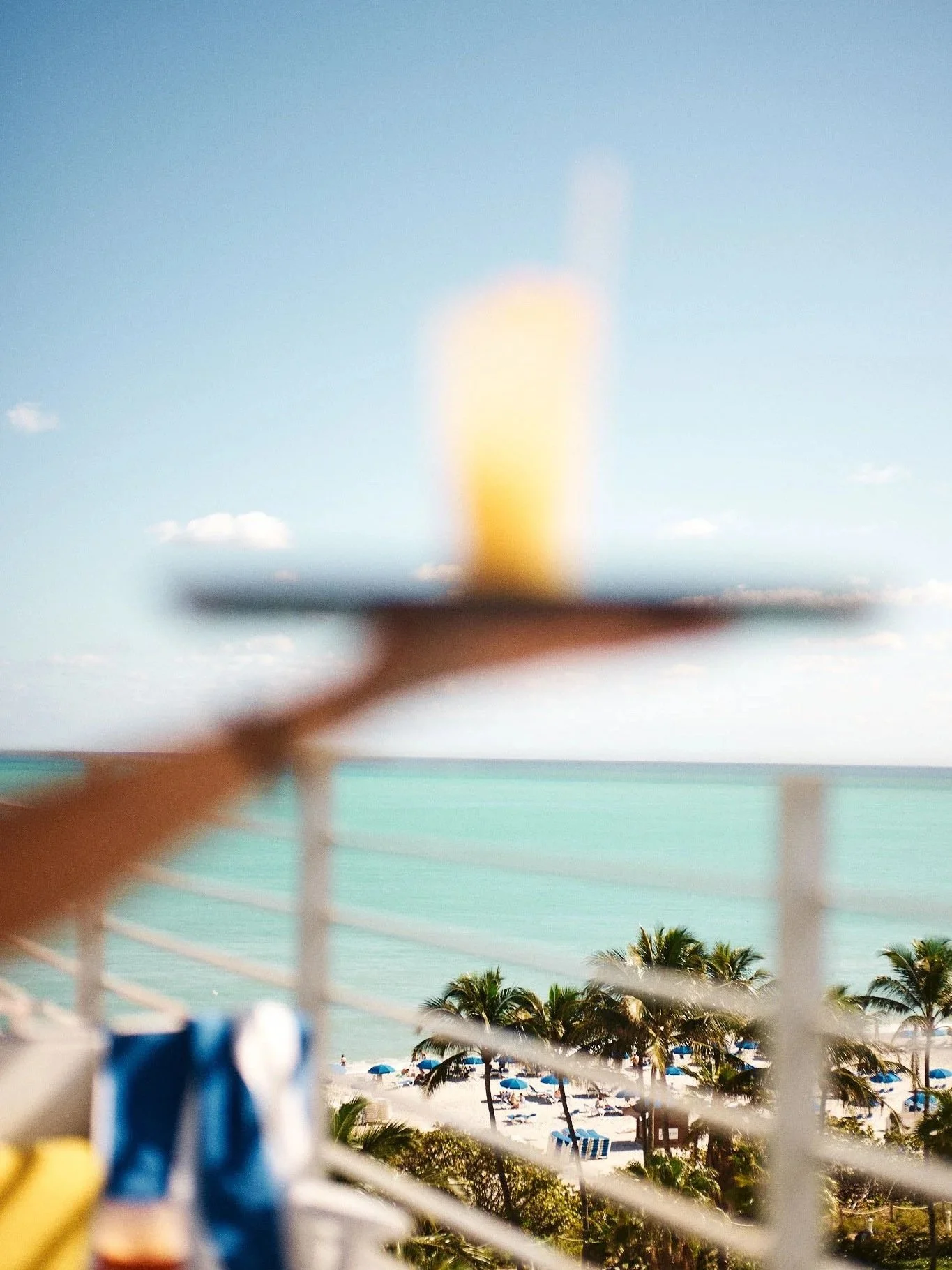 Blurred shot of a beach with palm trees, umbrellas, and the ocean in the background, with a person holding a tray with a yellow drink in the foreground.