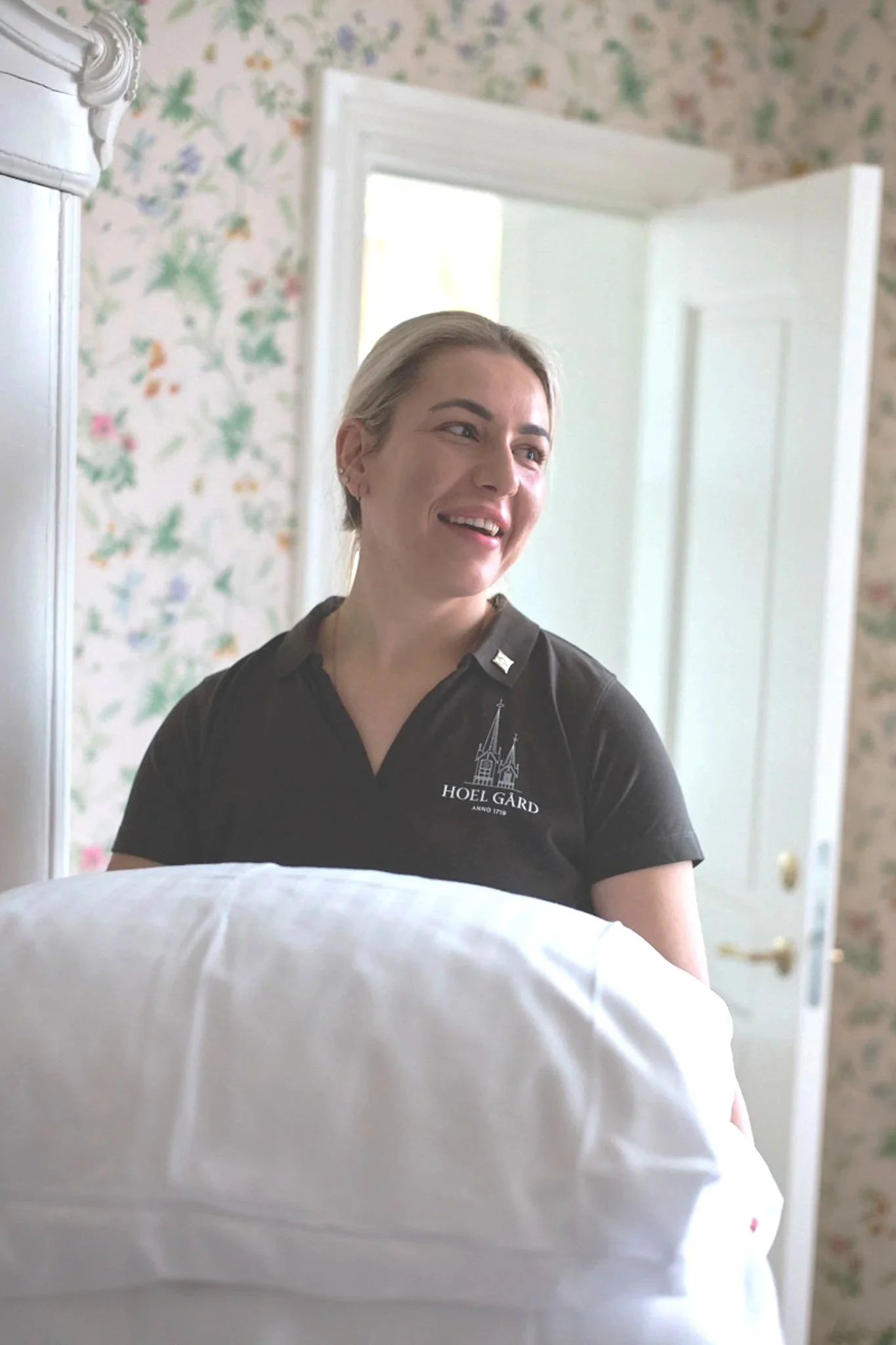 A woman in a black polo shirt with 'HOEL GARD' embroidered on it stands in a room with floral wallpaper, smiling as she looks to her right, with a bed or pillow partially visible in the foreground and a white door in the background.