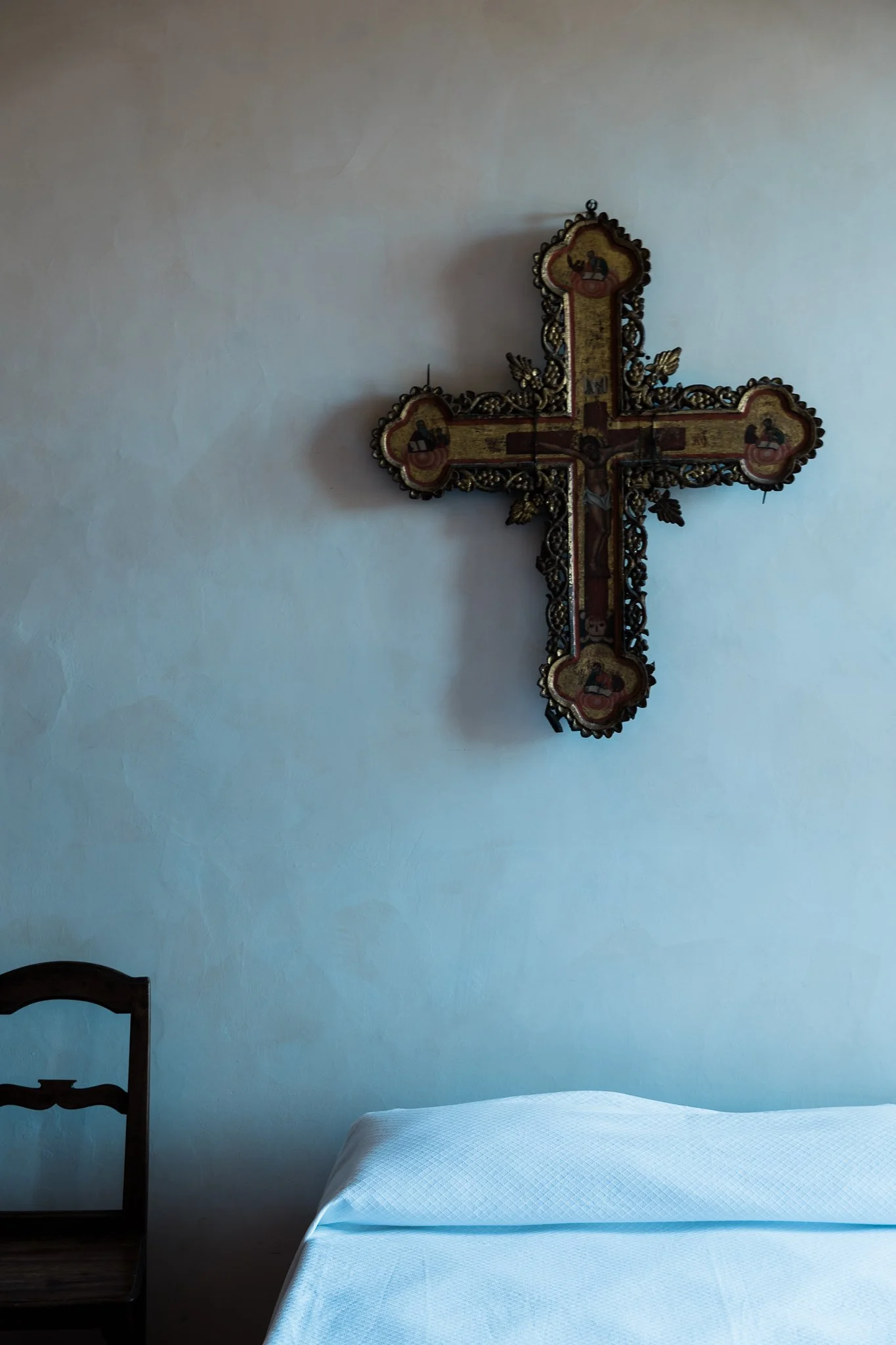 A wall with a crucifix and a small wooden chair next to a bed with a white bedspread.