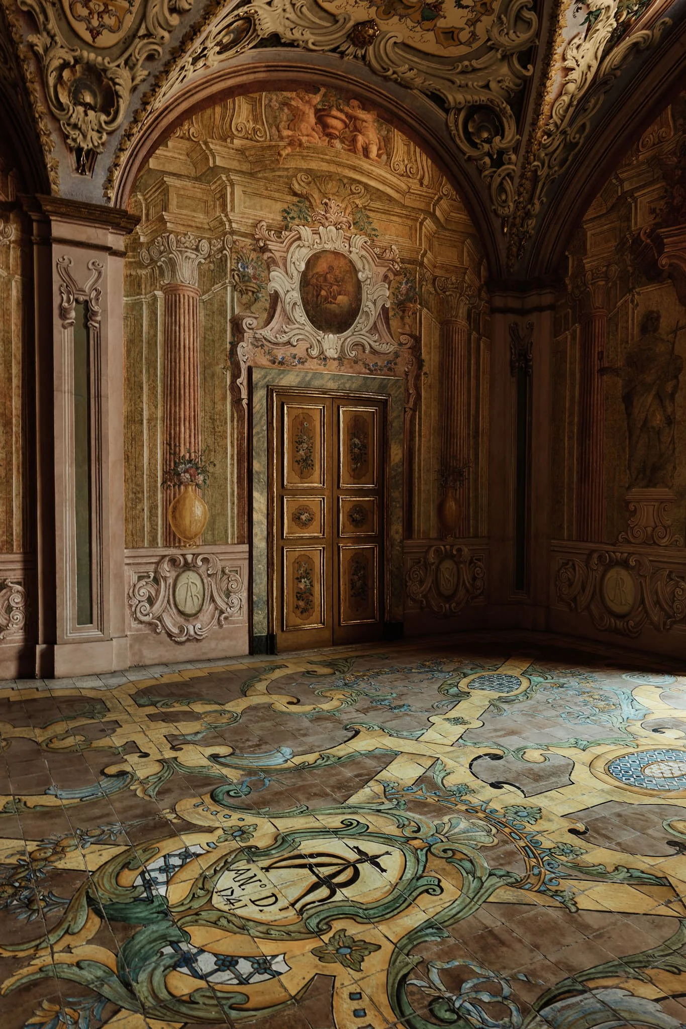 An ornately decorated interior room with intricate wall paintings, a wooden door, and a highly detailed, patterned floor.