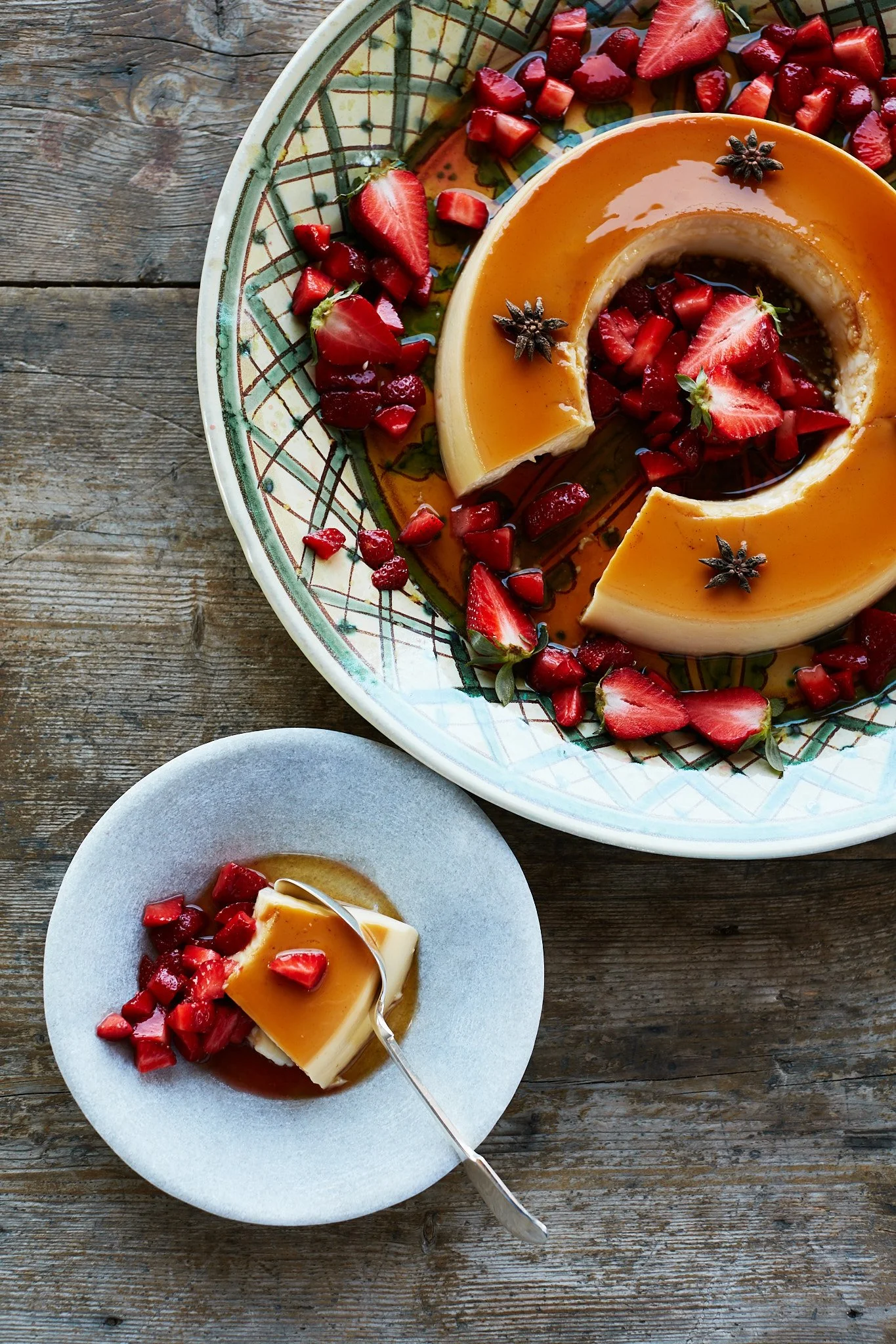 A caramel flan garnished with strawberries, chopped strawberries, star anise, served on a decorated plate. A slice of the flan on a small gray plate, with a fork, also topped with strawberries.