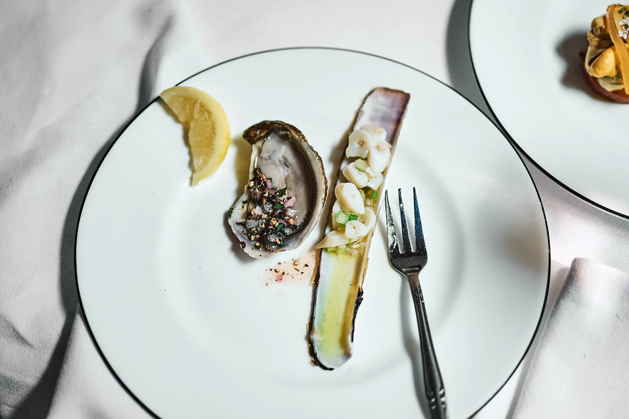 Empty white plate with a lemon wedge, half an oyster topped with spices, a slice of bread with chopped vegetables, and a fork, on a white tablecloth with another plate partially visible.