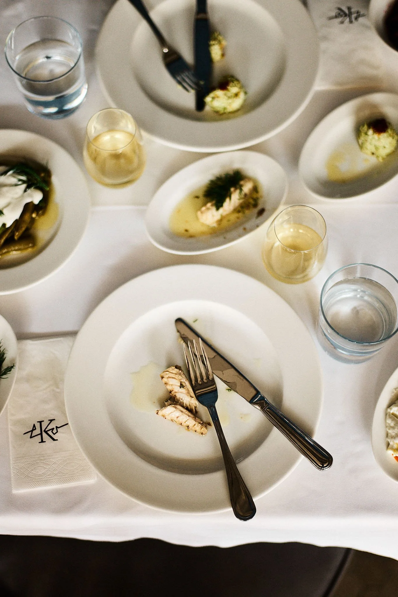 Empty white plate with a fork and knife, remnants of cooked fish, on a white tablecloth surrounded by glasses of water and wine, and several empty or partially eaten dishes with food, suggestive of a finished or nearly finished meal.