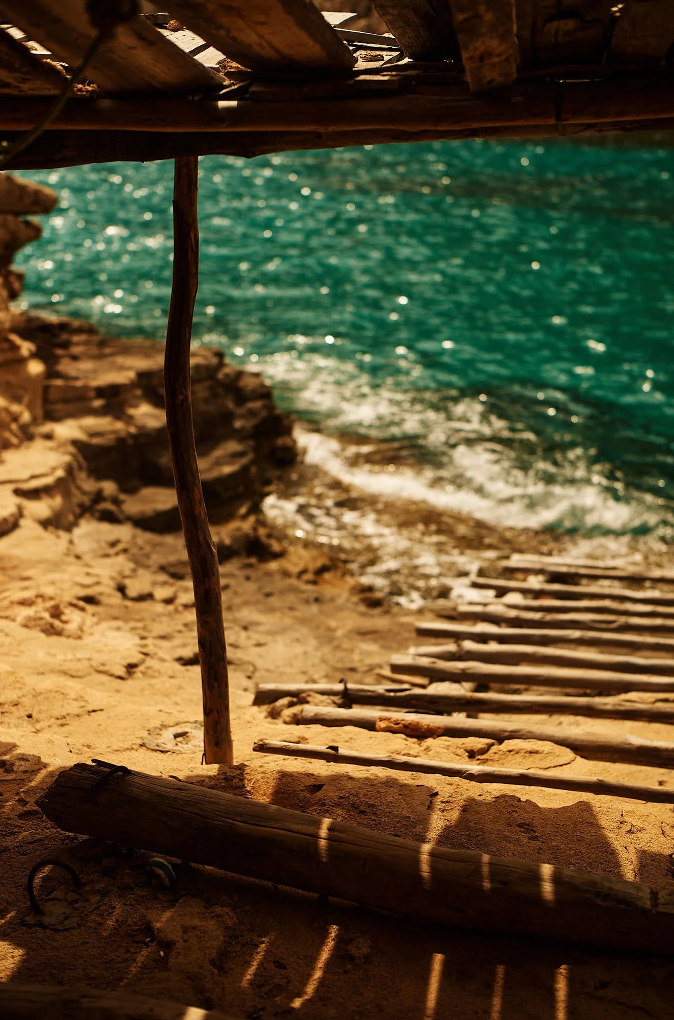 View of a sandy beach with wooden driftwood hut framing the turquoise ocean in the background, with waves gently hitting the shore.
