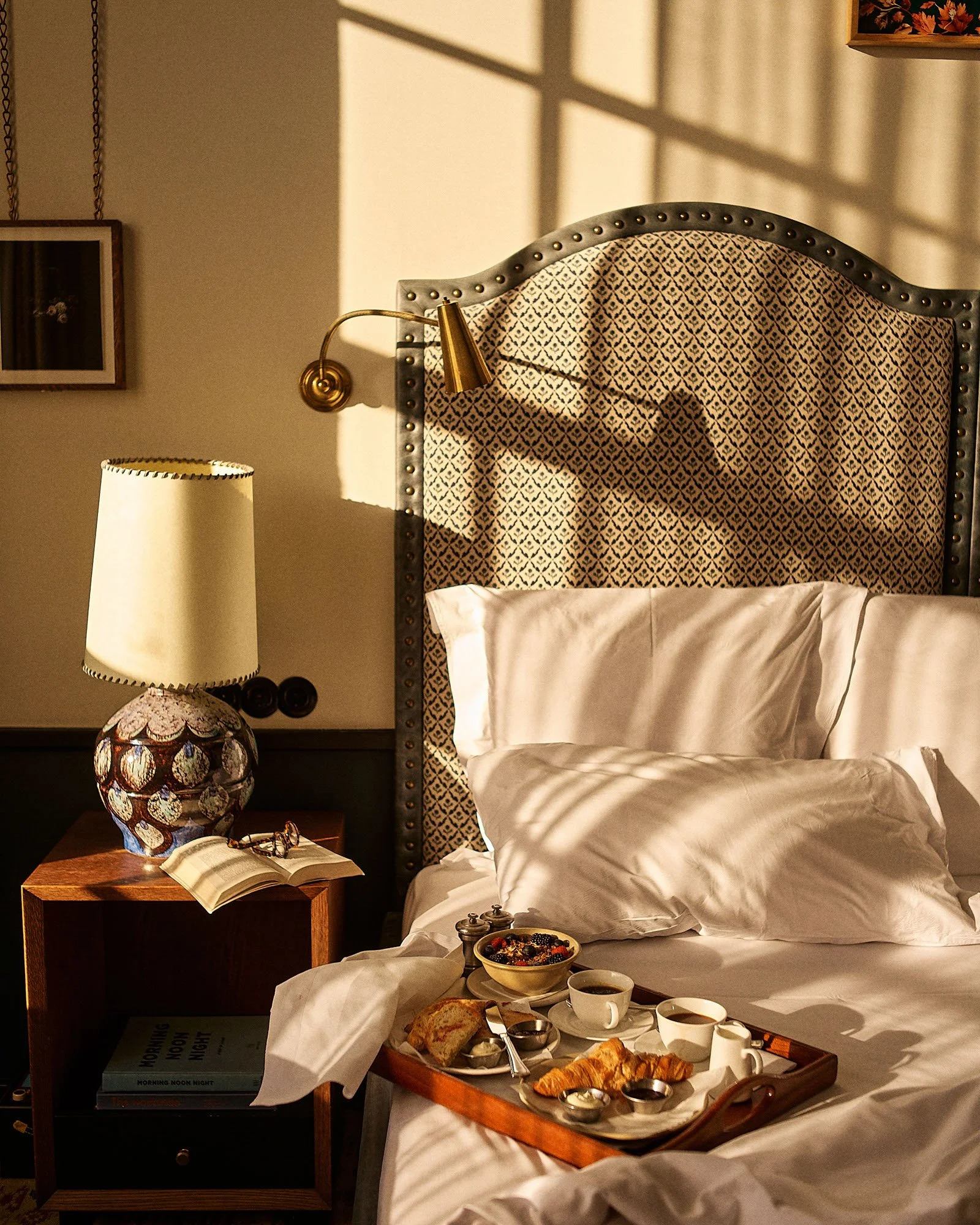 A cozy bedroom with sunlight casting shadows on a bed with white bedding, a wooden tray of breakfast items, a nightstand with a decorative lamp, an open book, and a patterned headboard.