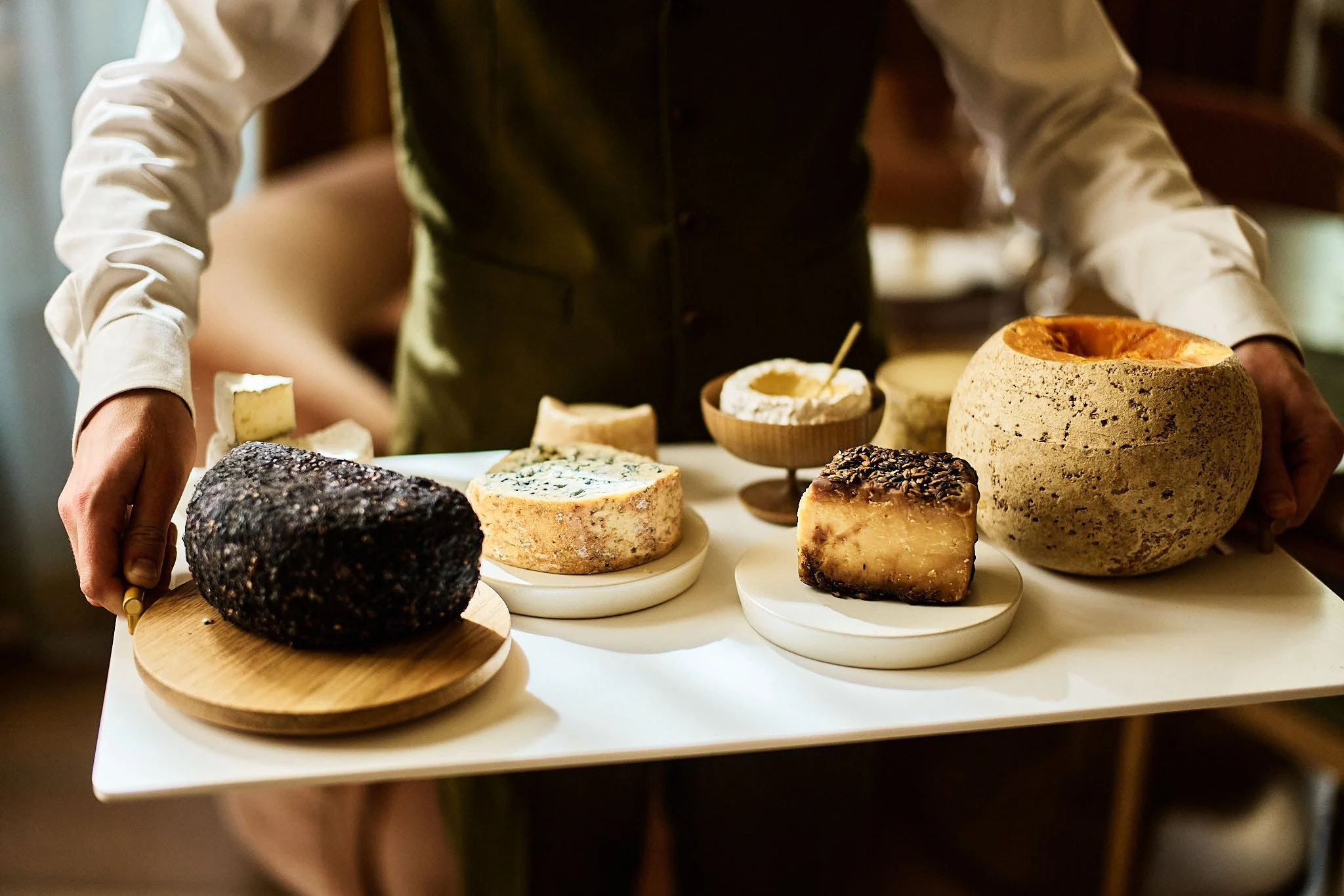 Person holding a white tray with various cheeses, including a large wheel of cheese, blue cheese, and other cheese blocks, with small bowls of accompaniments in the background.