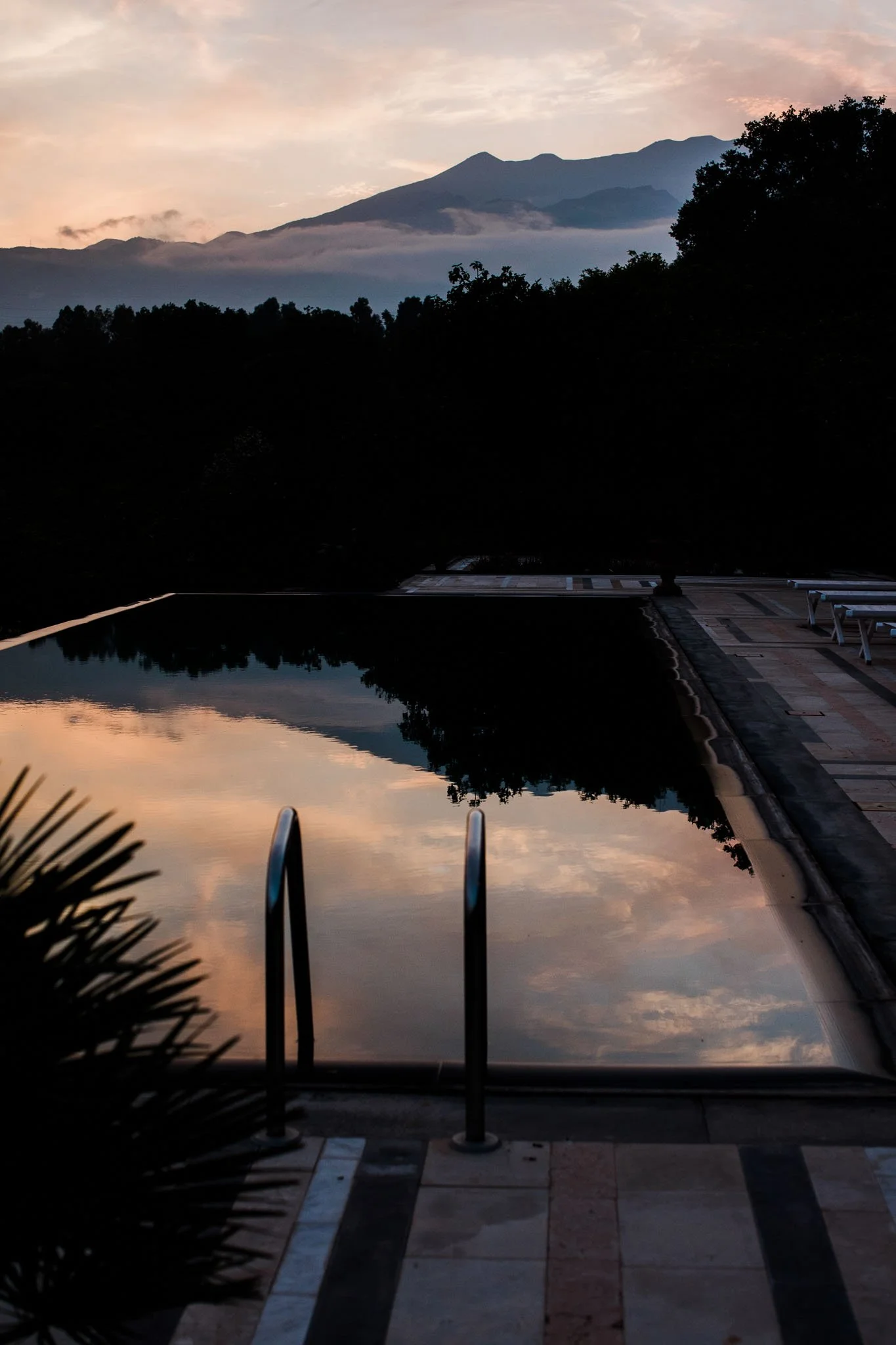 Reflection of the sky and mountains in an outdoor pool at sunset, with poolside chairs and surrounding greenery.