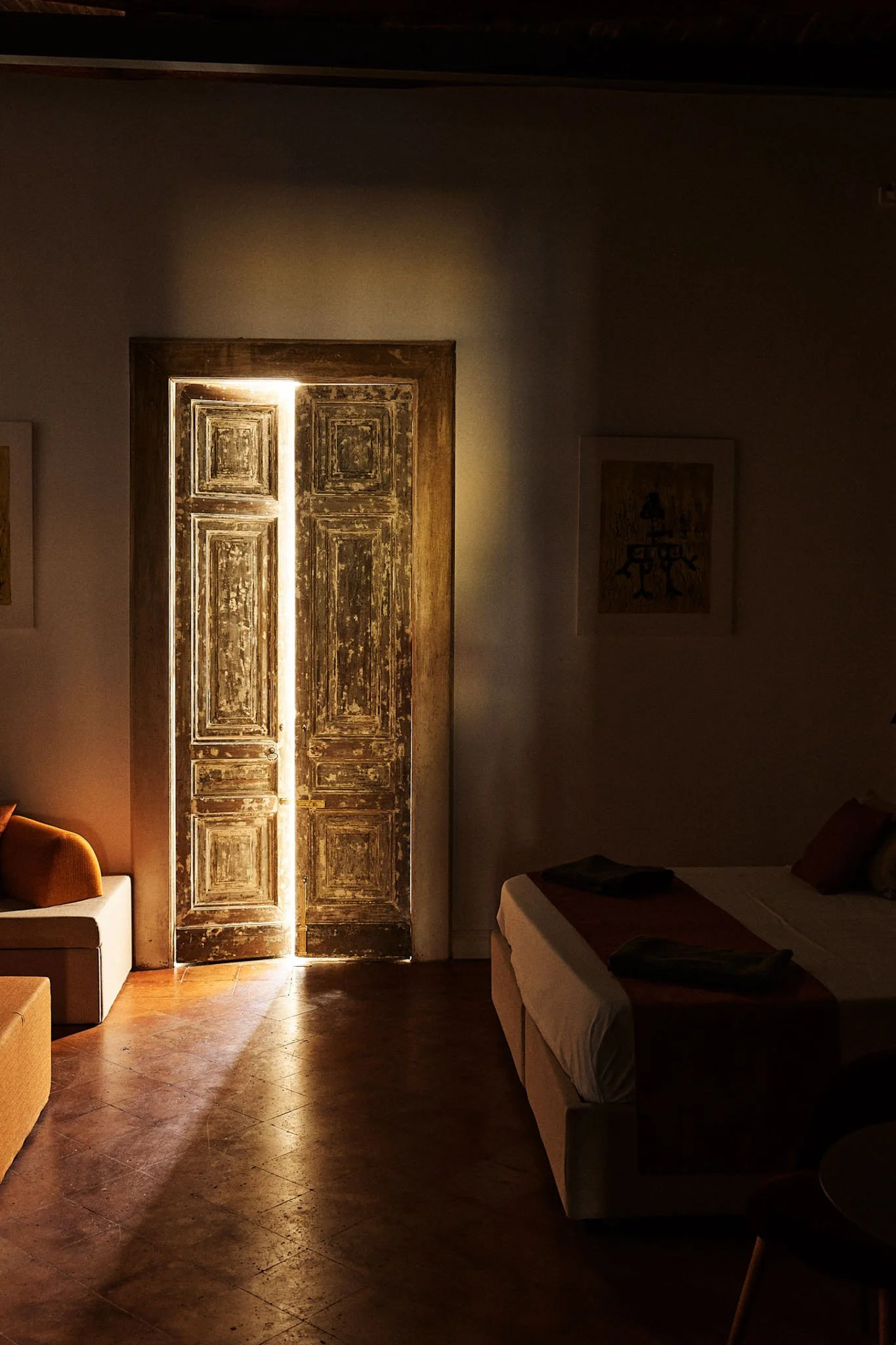 A dimly lit room with a wooden double door slightly ajar, allowing a narrow beam of sunlight to shine through, illuminating part of the floor and the edge of a bed. There is a chair partially visible on the right and framed artwork on the wall.