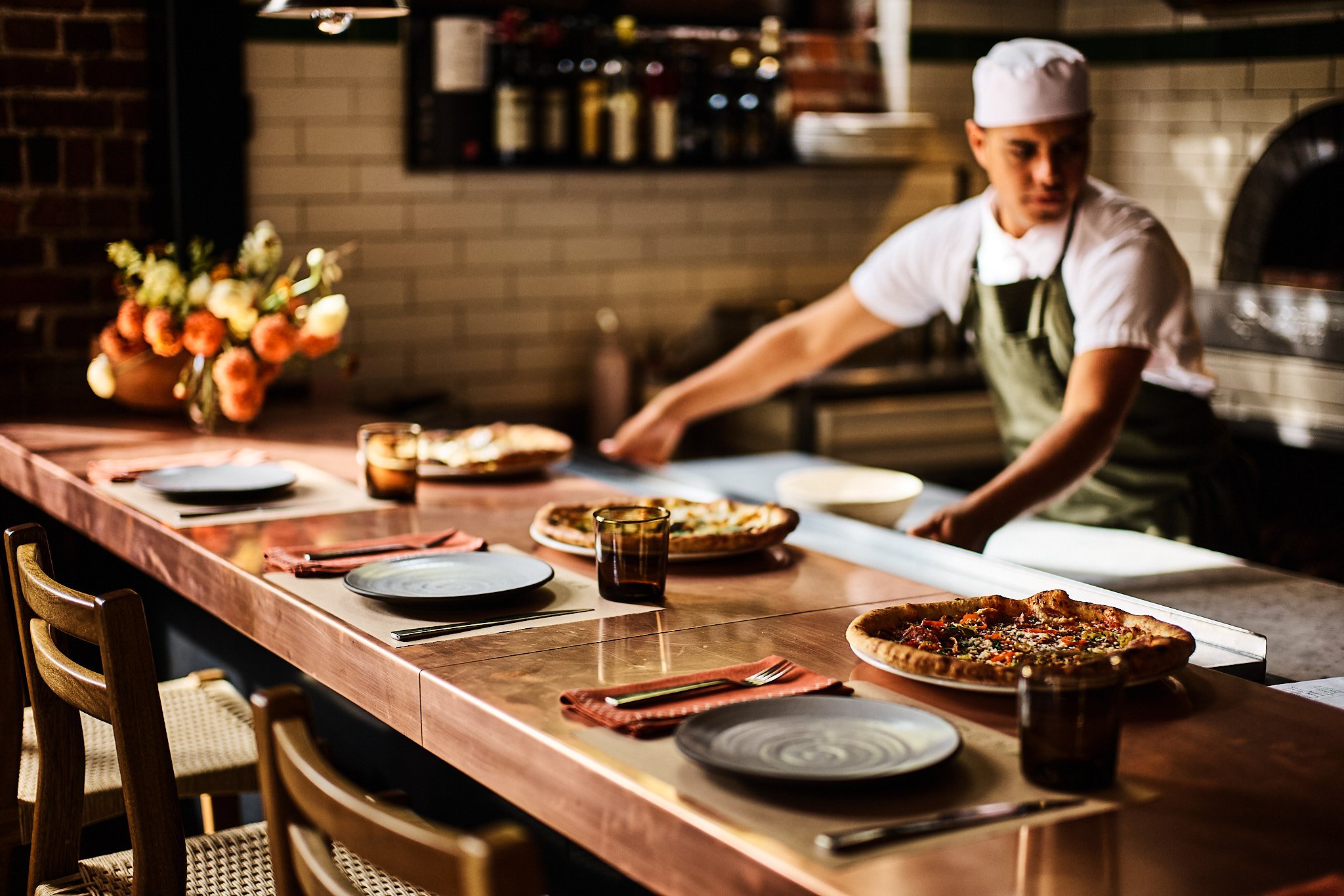 A person in a chef's hat and apron preparing pizzas behind a dining counter in a restaurant with a brick wall and shelves in the background. The table has plates, drink glasses, napkins, and several pizzas.