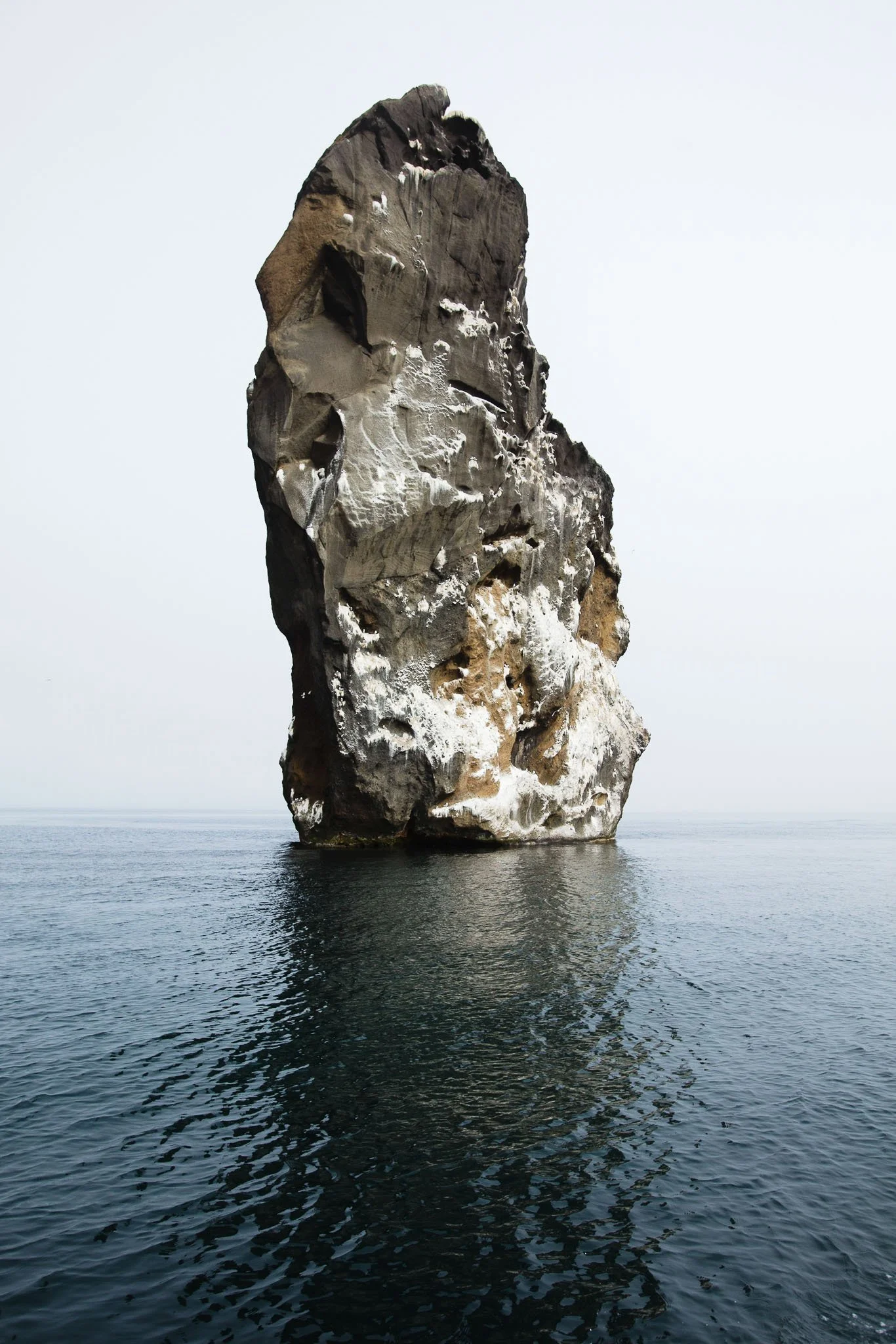 Large solitary sea stack with a rugged surface partially covered in ice, rising from calm water under a cloudy sky.