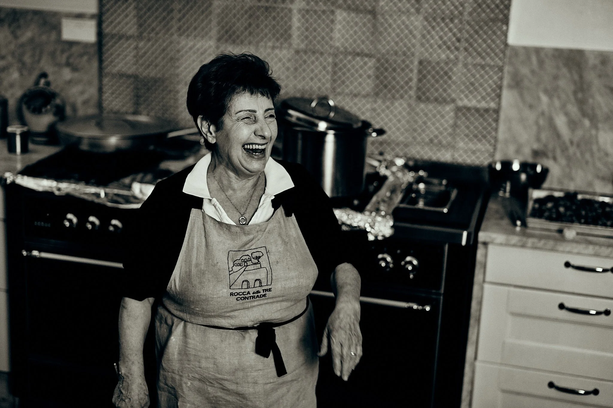 A smiling elderly woman wearing an apron in a kitchen, standing in front of a stove with pots and kitchen utensils.