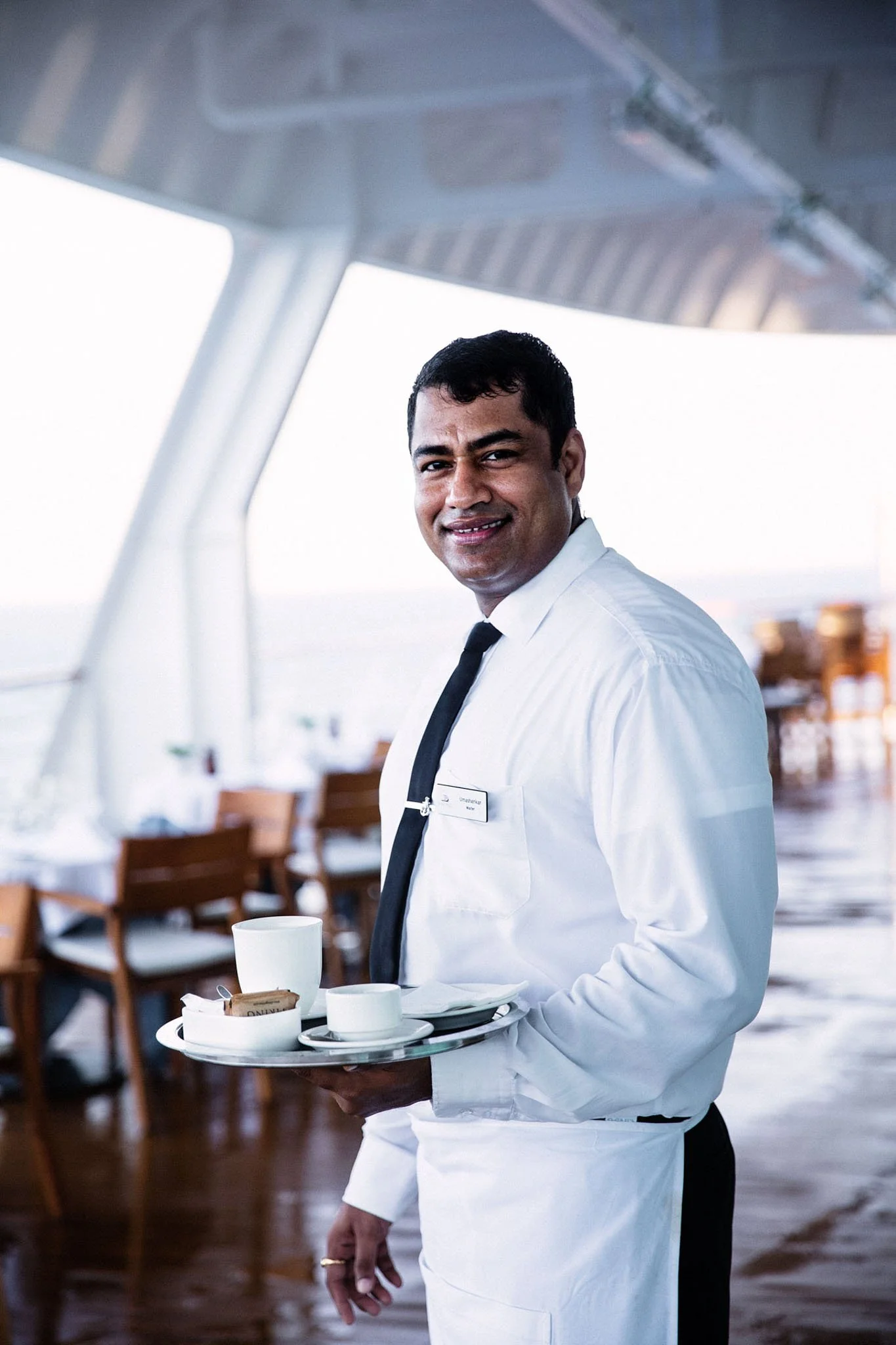 A smiling waiter in a white shirt and black tie holding a tray with cups and tea bags inside a ship's dining area.