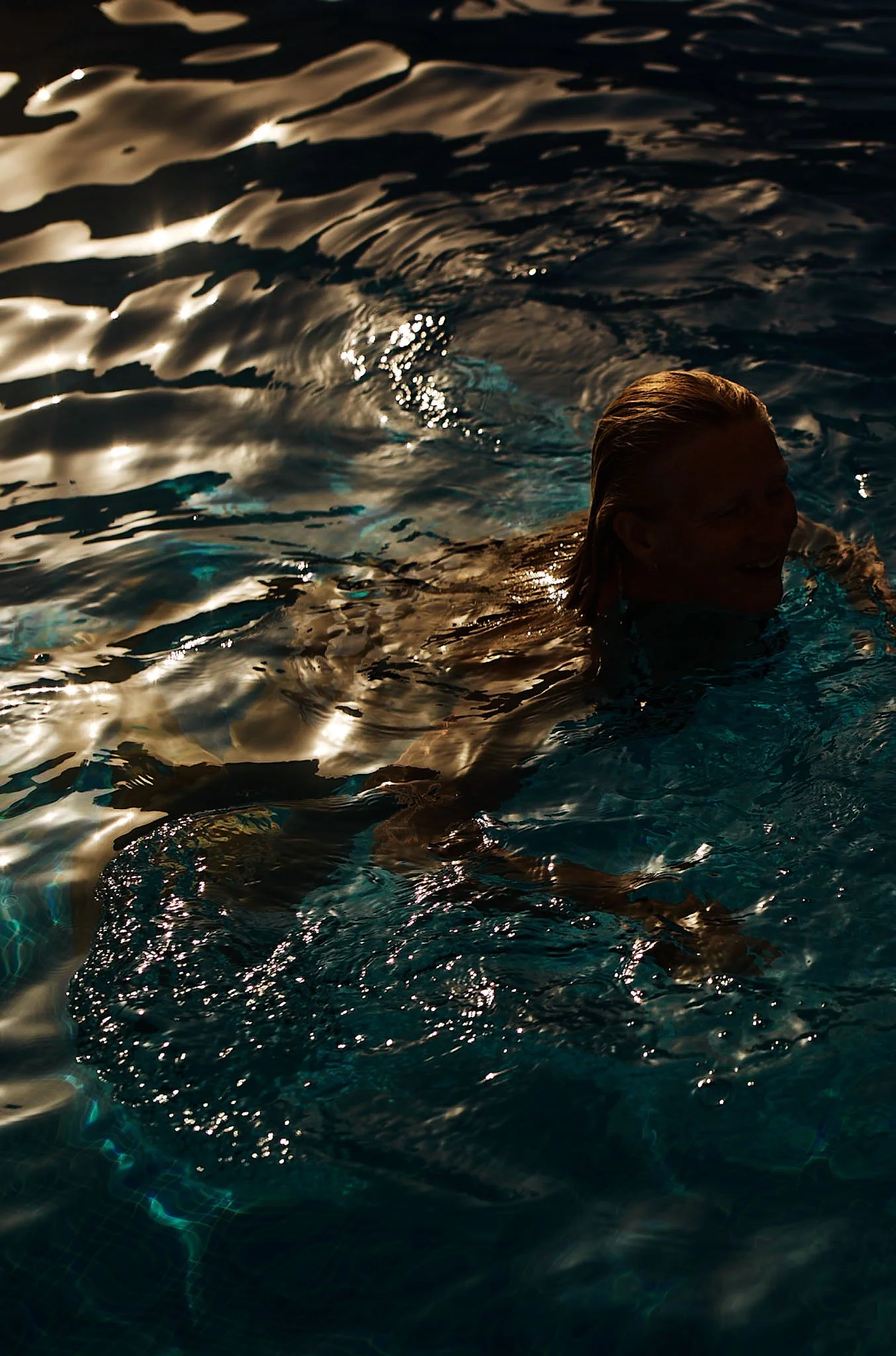 Person swimming in a pool during sunset or sunrise, with reflections on the water surface.
