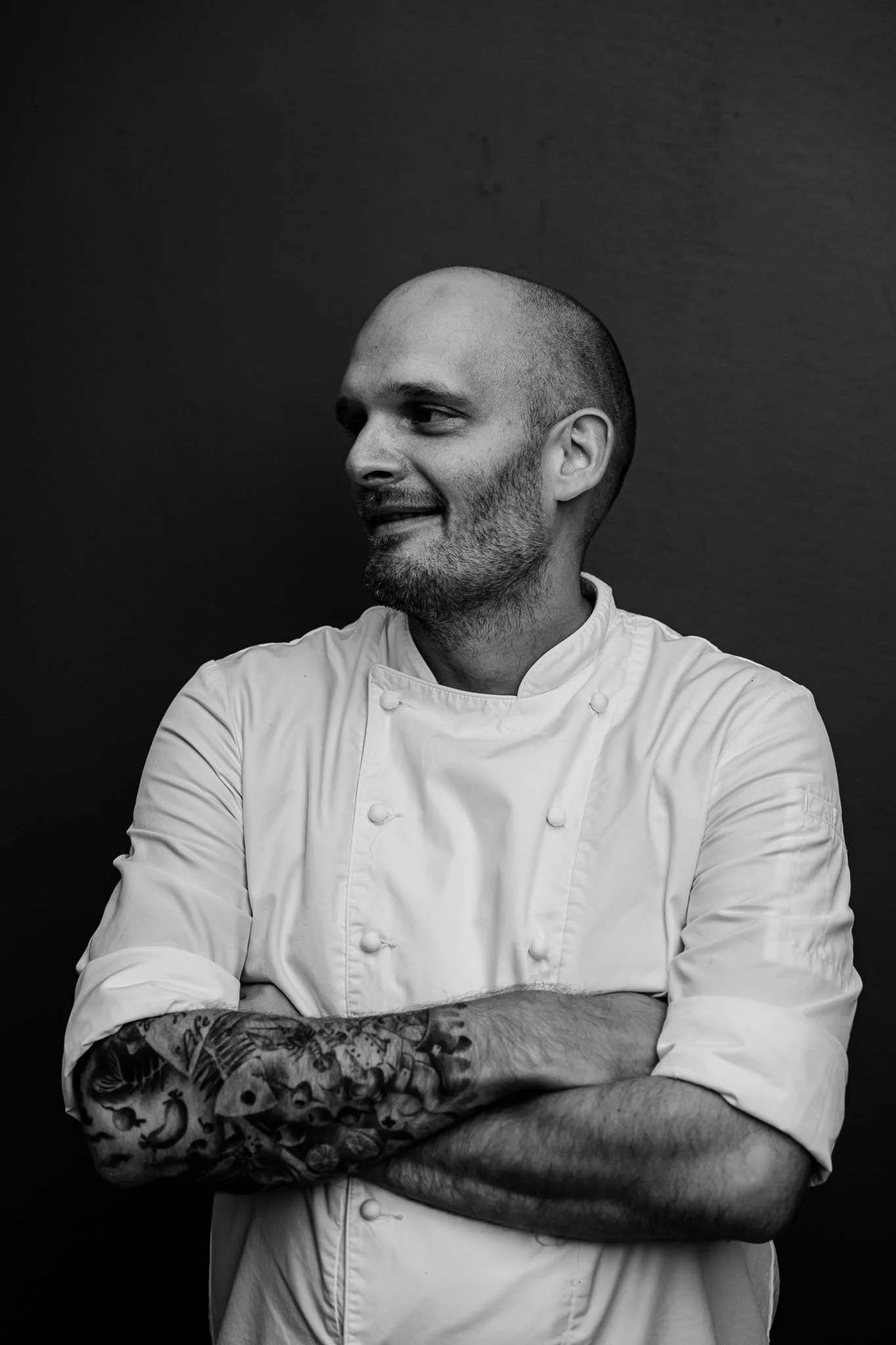 Black and white photo of a male chef with a tattooed arm, wearing a chef's coat, standing against a dark background with arms crossed.