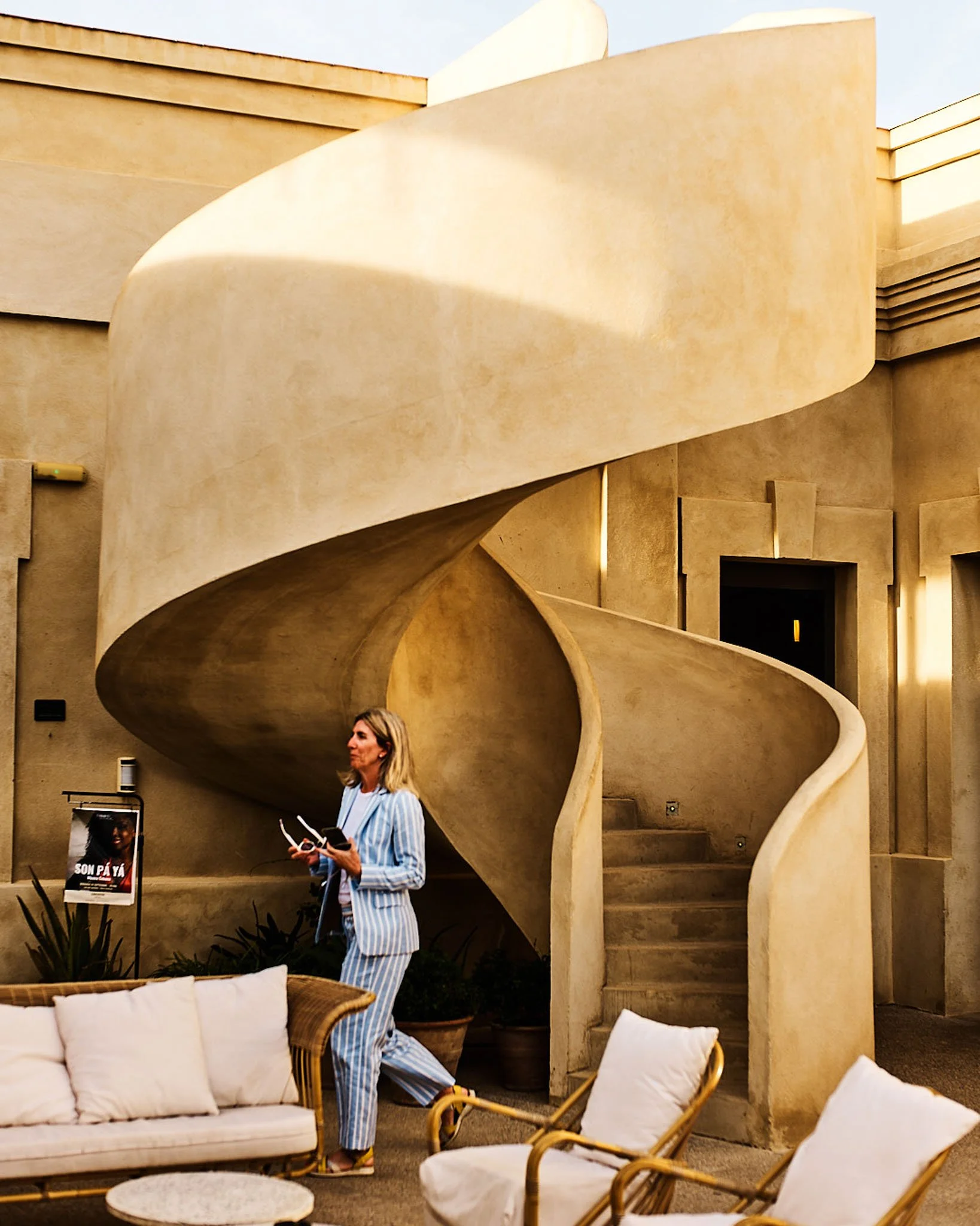 Woman in striped pajamas holding a drone and a phone, standing beneath an outdoor spiral staircase, with lounge chairs and potted plants in the foreground.