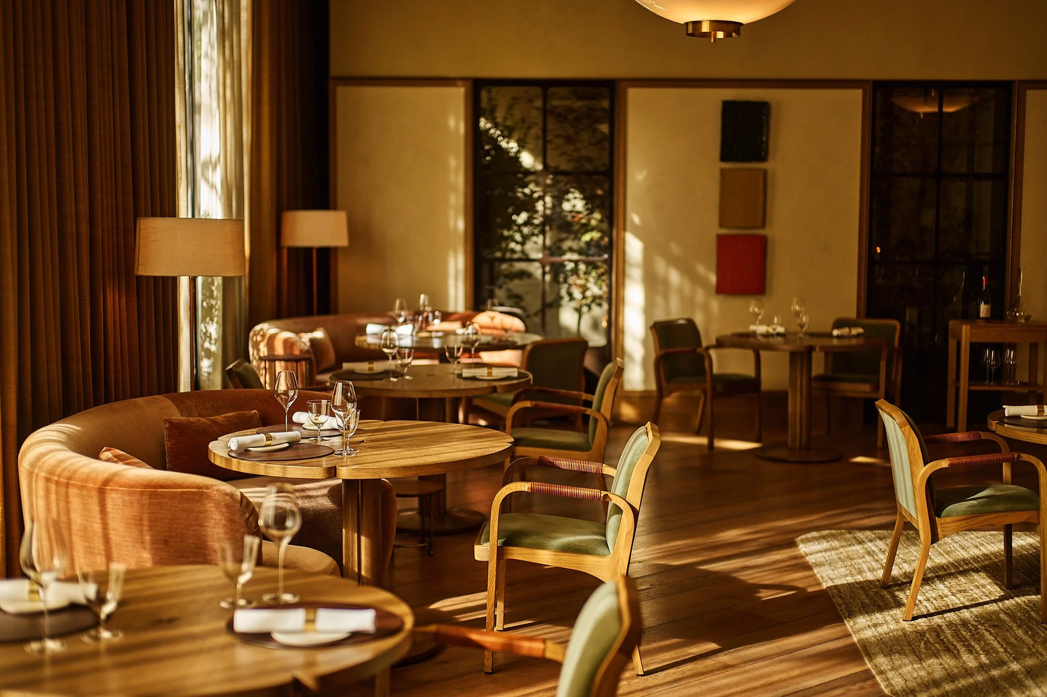 A warmly lit restaurant with wooden tables and chairs, set with wine glasses and napkins, featuring large windows with curtains and sunlight filtering through, creating a cozy atmosphere.