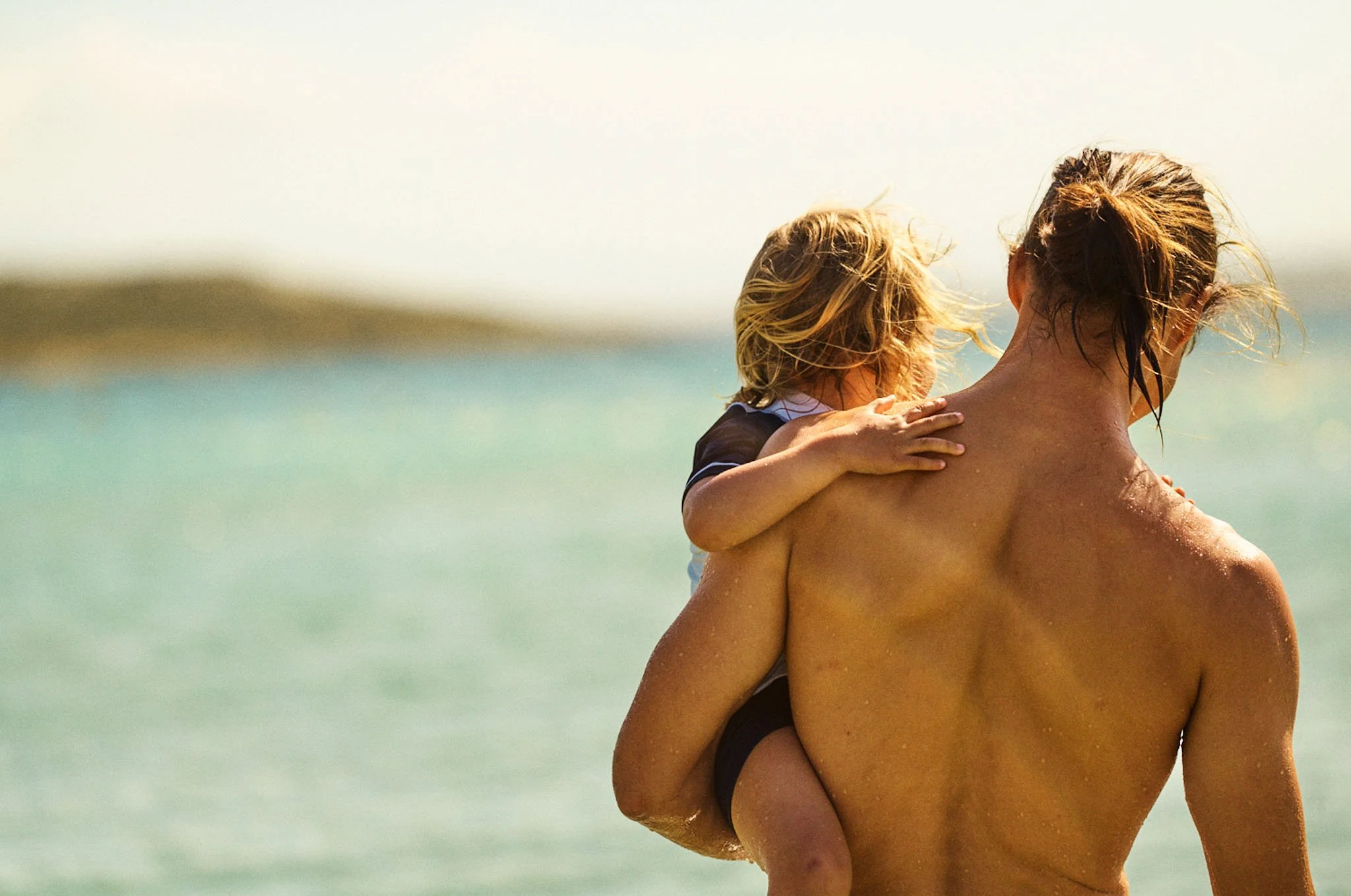 A woman carrying a young child on her back at the beach with water and distant land in the background.