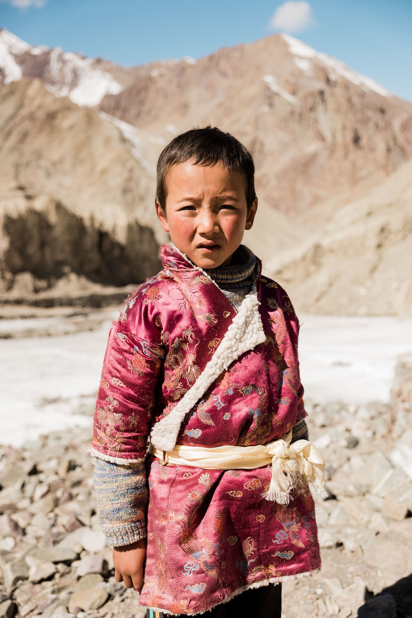 A young boy standing outdoors in a mountainous landscape, wearing a traditional red silk garment with intricate embroidery, a cream-colored sash, and a warm sweater underneath.